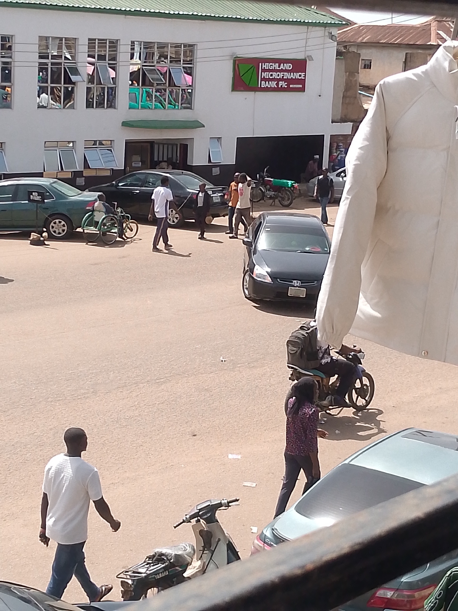 The image depicts a street scene outside a bank in Bukuru, Nigeria. Several people are visible, including a man in a white t-shirt and blue jeans walking towards the right, and another person in a patterned shirt and dark pants walking away from the viewer. A person in a wheelchair is also present, being pushed by another individual.
Vehicles are a prominent feature of the scene. A green sedan is parked on the left, with a sign on its windshield that is too small to read. A black sedan is in the middle of the road, and a motorcycle is parked near a building on the right. A person is riding another motorcycle, carrying a backpack.
The setting appears to be daytime, with bright sunlight casting shadows. The architecture of the bank building, with its white facade and multiple windows, suggests an urban environment. A sign on the bank reads "HIGHLAND MICROFINANCE BANK Plc".
The overall atmosphere seems to be one of everyday activity, with people going about their business. There are no overt indications of any particular event or strong emotions. The weather appears to be clear and sunny.