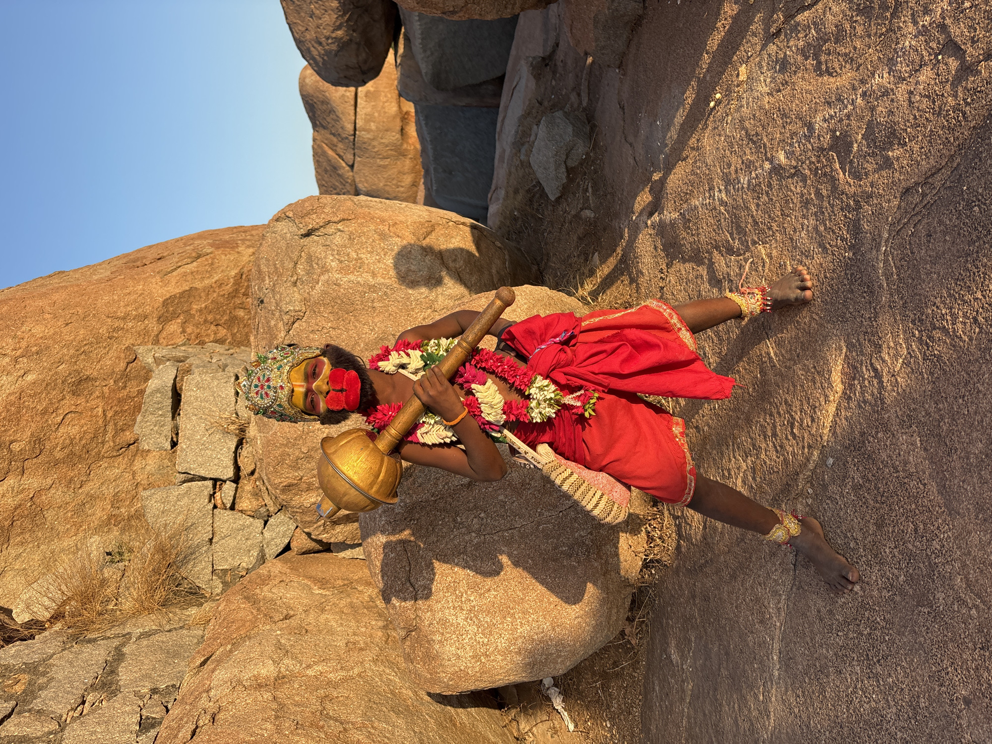A color photograph depicts a person dressed as the Hindu deity Hanuman, situated amongst large rock formations, likely in Kamalapura, India. The individual is adorned with a headdress, facial paint including a red mouth, necklaces of flowers, and bracelets. They are clad in red clothing and holding a golden mace. The setting appears to be a natural landscape with smooth, weathered rocks and a clear sky in the background.