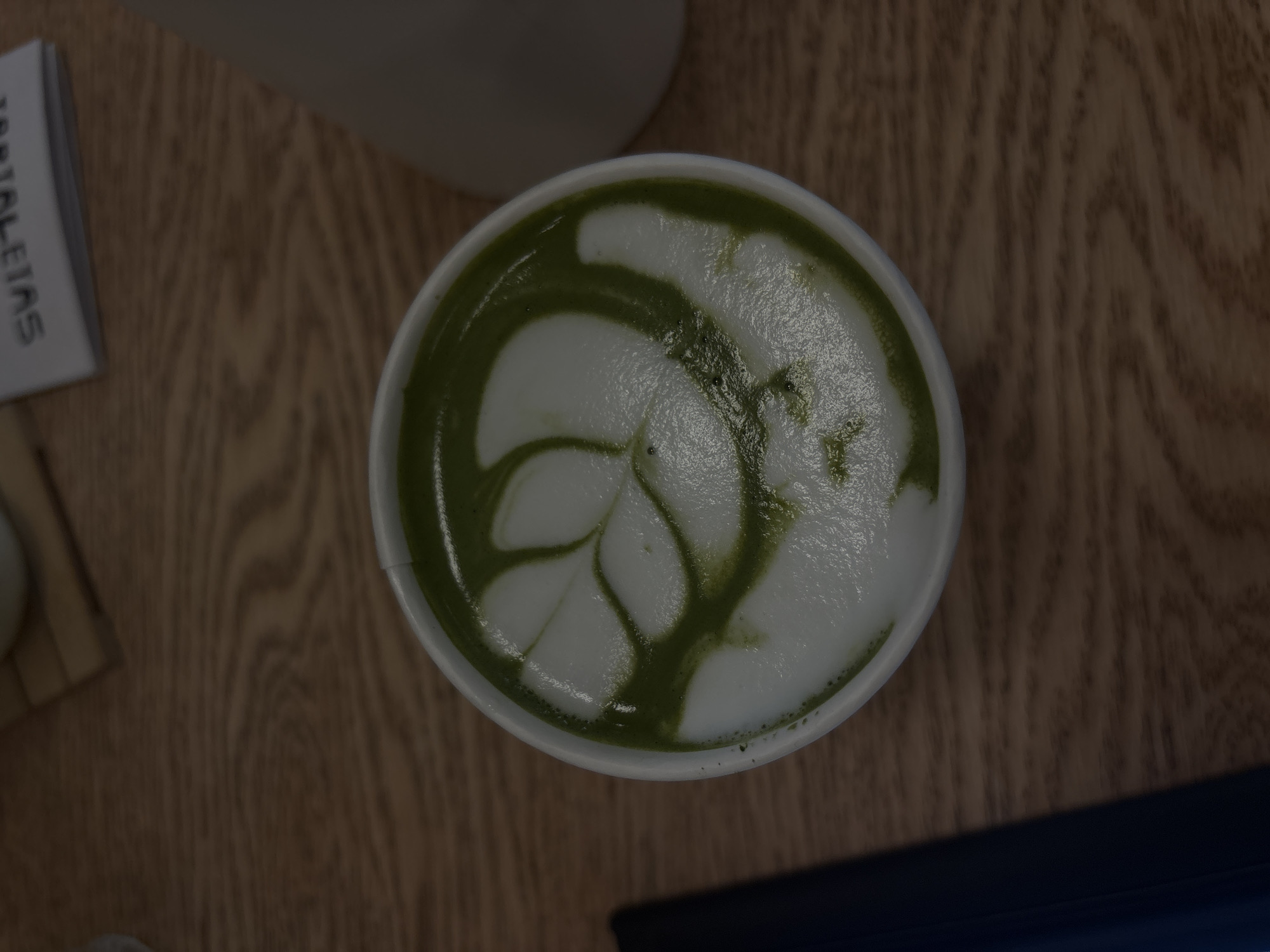 The image shows a top-down view of a cup of matcha latte with latte art resembling a stylized leaf or floral design. The beverage sits on a wooden surface. Part of a white container is visible in the upper center of the image, slightly out of focus. A small box with the word "TARTALEITAS" printed on it is visible on the left edge of the frame. A dark object (possibly a laptop or tablet) is partly visible in the lower right corner of the image. The location is not discernible from the image alone, and there's no evidence to suggest it's in Puebla, Mexico.