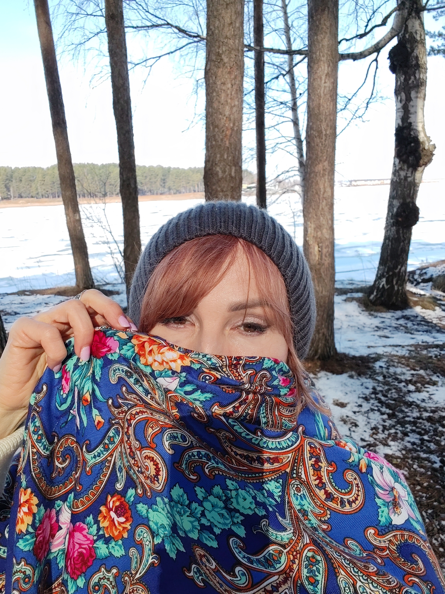 A woman is standing outdoors in a winter landscape. She has a gray beanie on her head and her reddish-brown hair is visible at the front. Her eyes are visible above a brightly colored patterned scarf that she holds up to cover the lower part of her face. The scarf has a blue background with paisley and floral designs in shades of pink, orange, and green. The woman's hand is visible holding the edge of the scarf; her fingernails are painted pink. In the background are several trees with bare branches, and beyond them, a partially frozen body of water with a forest line on the opposite shore. The ground is covered with patches of snow and dried leaves. The location is likely Chaikovsky, Russia, given the environment and typical Russian clothing.