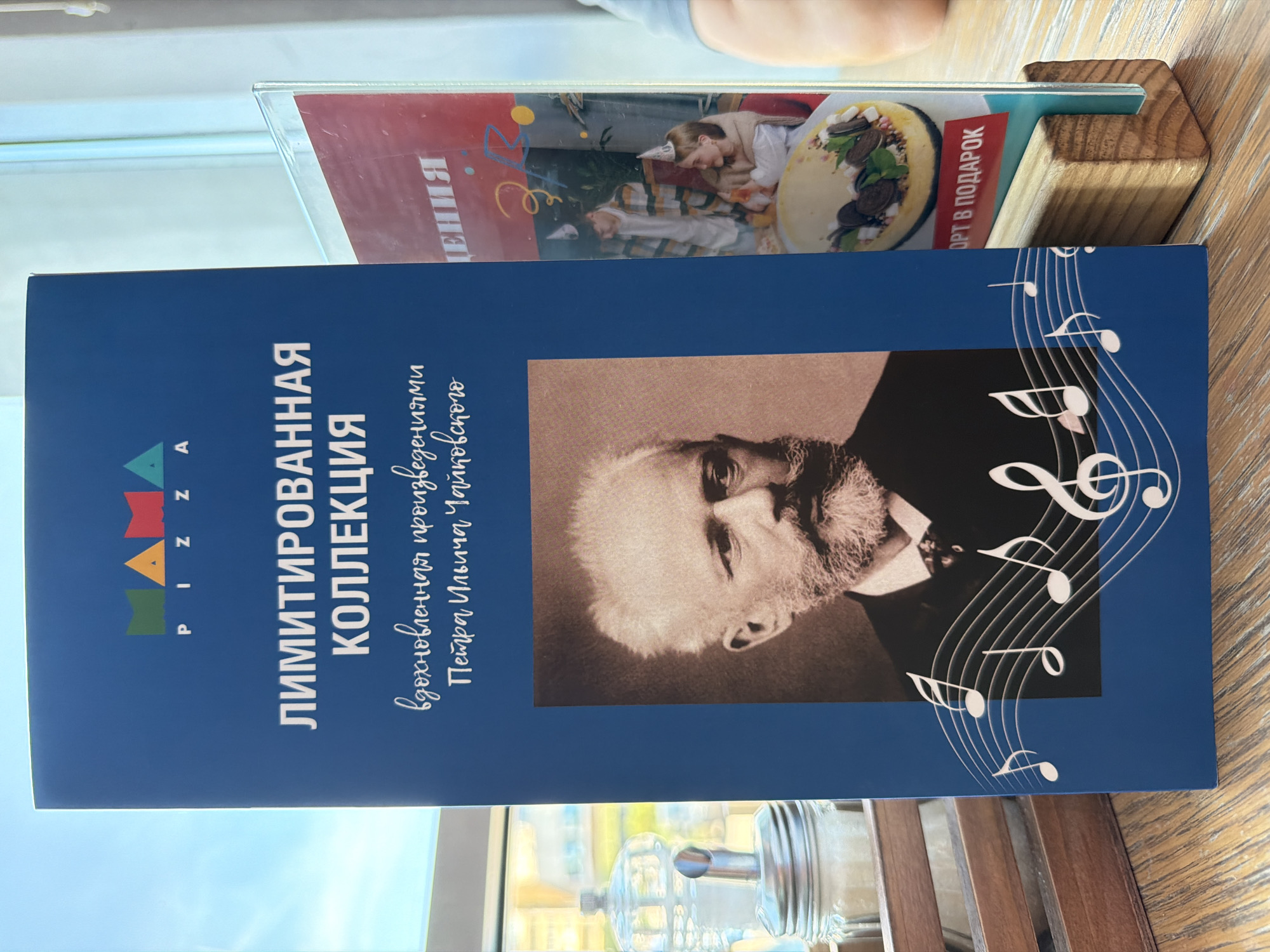 The image shows a blue menu for "Mama Pizza" with a collection inspired by Tchaikovsky. A sepia-toned portrait of Pyotr Ilyich Tchaikovsky is displayed prominently. Musical notes are graphically illustrated on the menu. To the right, a clear plastic menu holder displays another menu with a picture of a cheesecake and people wearing party hats. The menus are placed on a wooden surface, possibly a table, in what appears to be a restaurant setting. A glass sugar dispenser is visible in the background on the left, suggesting a cafe or dining area. The scene is illuminated by natural light coming from a window. There are no discernible landmarks or visual cues that would allow for specific location identification in Izhevsk, Russia.