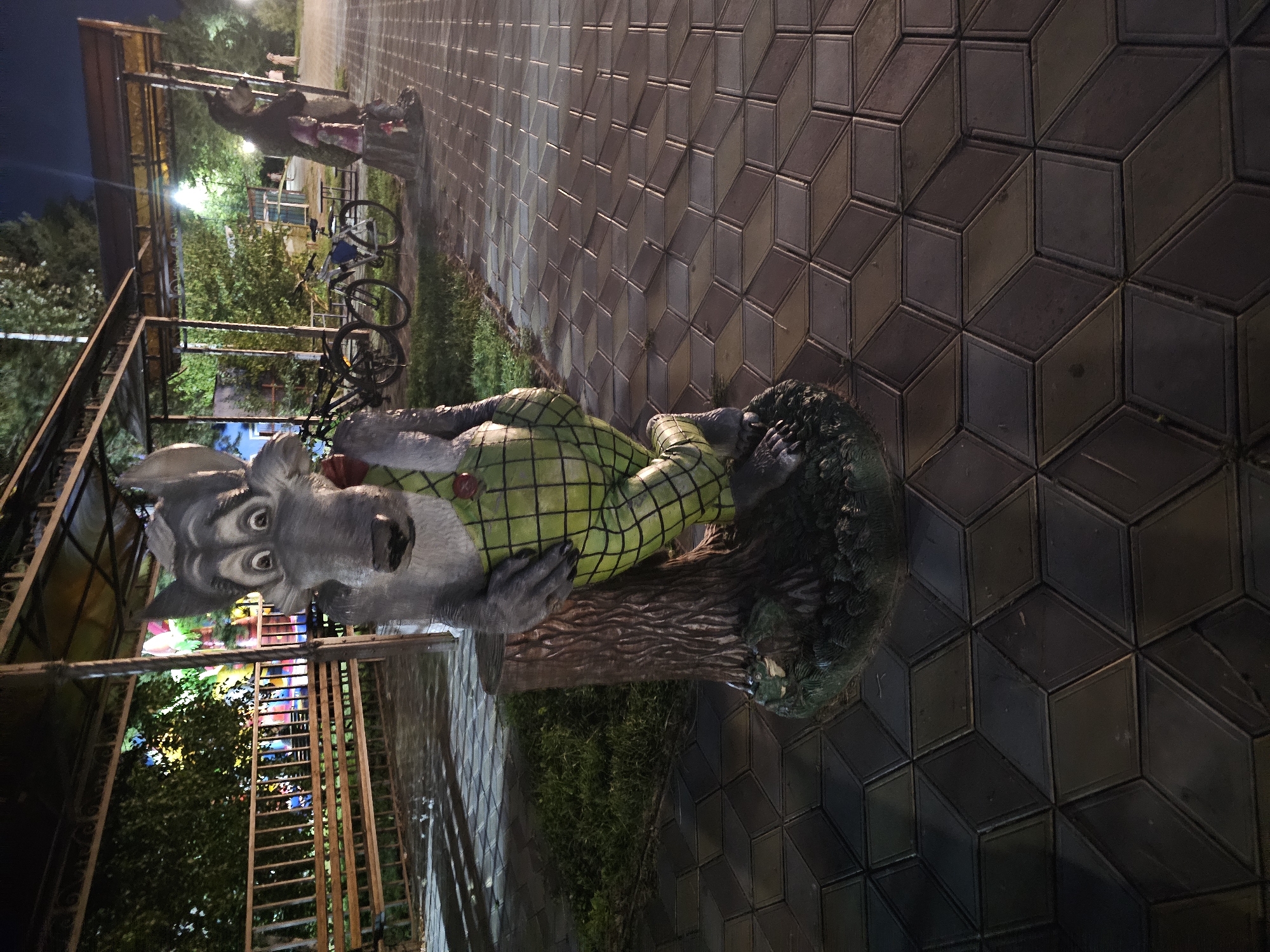 This image shows a statue of a cartoon wolf character dressed in overalls and standing next to a tree stump. The statue is located in an outdoor park area with paved walkways. In the background, there are benches, trees, bicycles, and some amusement park rides. The lighting suggests it is nighttime. This scene is located at the Central Park in Cherkessk, Russia.
