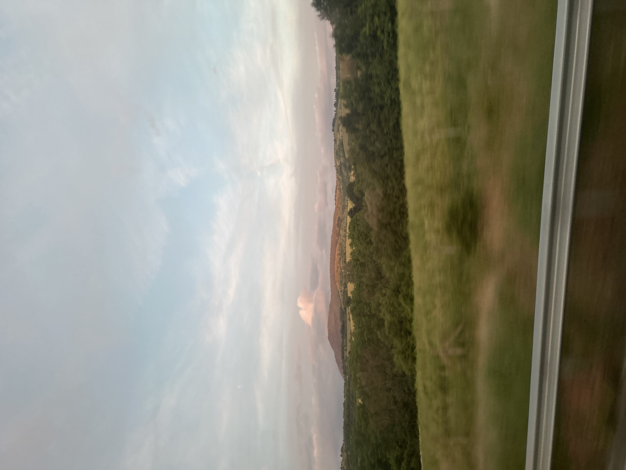 An exterior, eye-level, medium shot captures a rolling landscape under a pale, cloudy sky, likely during sunset, judging by the soft pink and orange hues that tint the clouds and highlight the distant hills. The foreground is dominated by a blurred, green grassy field, suggesting motion, possibly from a vehicle. A grey metal guardrail runs horizontally across the bottom of the frame. Behind the field, a dense line of dark green trees forms a treeline, with lighter green rolling hills and more distant, rounded mountains visible beyond them. A sliver of the moon is faintly visible in the upper left quadrant of the sky. The location is described as Osikovitsa, Bulgaria.