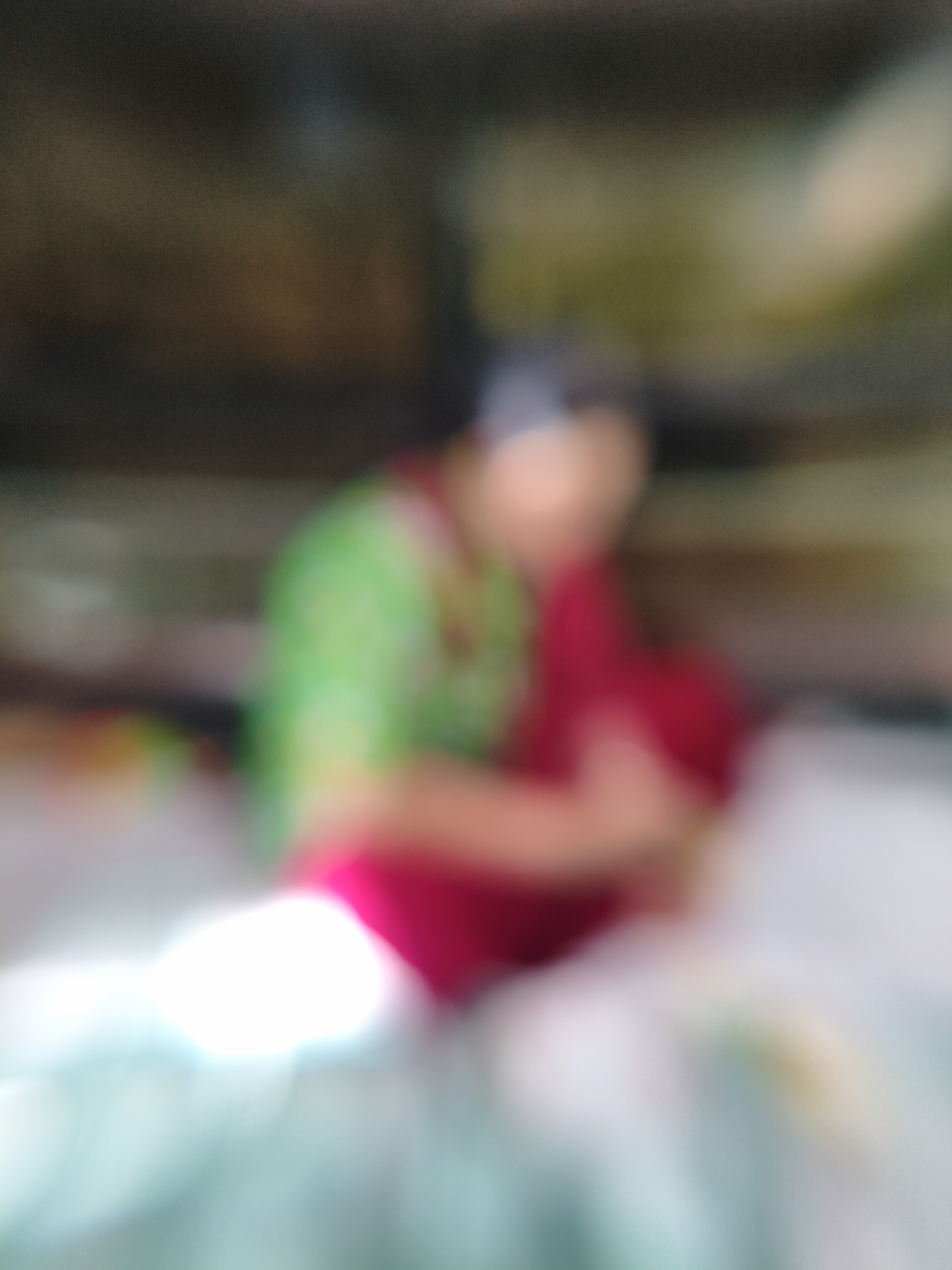 This media file is an extremely blurred and out-of-focus image, predominantly featuring a person as the central subject. The heavy blur gives the image an abstract quality.
The primary subject, likely a person, is situated in the mid-ground. They appear to be wearing a vibrant green upper garment and a striking red or magenta lower garment, possibly a skirt or draped fabric, with a darker head covering or hair. The person seems to be in a sitting or crouched position, with limbs indistinctly folded towards the body.
The background is uniformly dark and out of focus, showing indistinct shades of brown and green, suggesting a shadowed or interior setting. Hints of brighter, diffused light are visible in the upper right background.
The foreground features a significant area of intense brightness, appearing as a luminous white and light blue/teal blur, possibly indicating a strong light source, a highly reflective surface, or a light-colored object/fabric close to the camera.
Due to the extreme blur, no specific activity or event can be discerned, nor are any emotions visible on the person's face. The time of day or weather conditions are not apparent. There are no visible location cues within the image itself, and no text is present. The overall impression is one of soft colors and diffused light, with the bright clothing of the subject standing out against the darker background and luminous foreground.