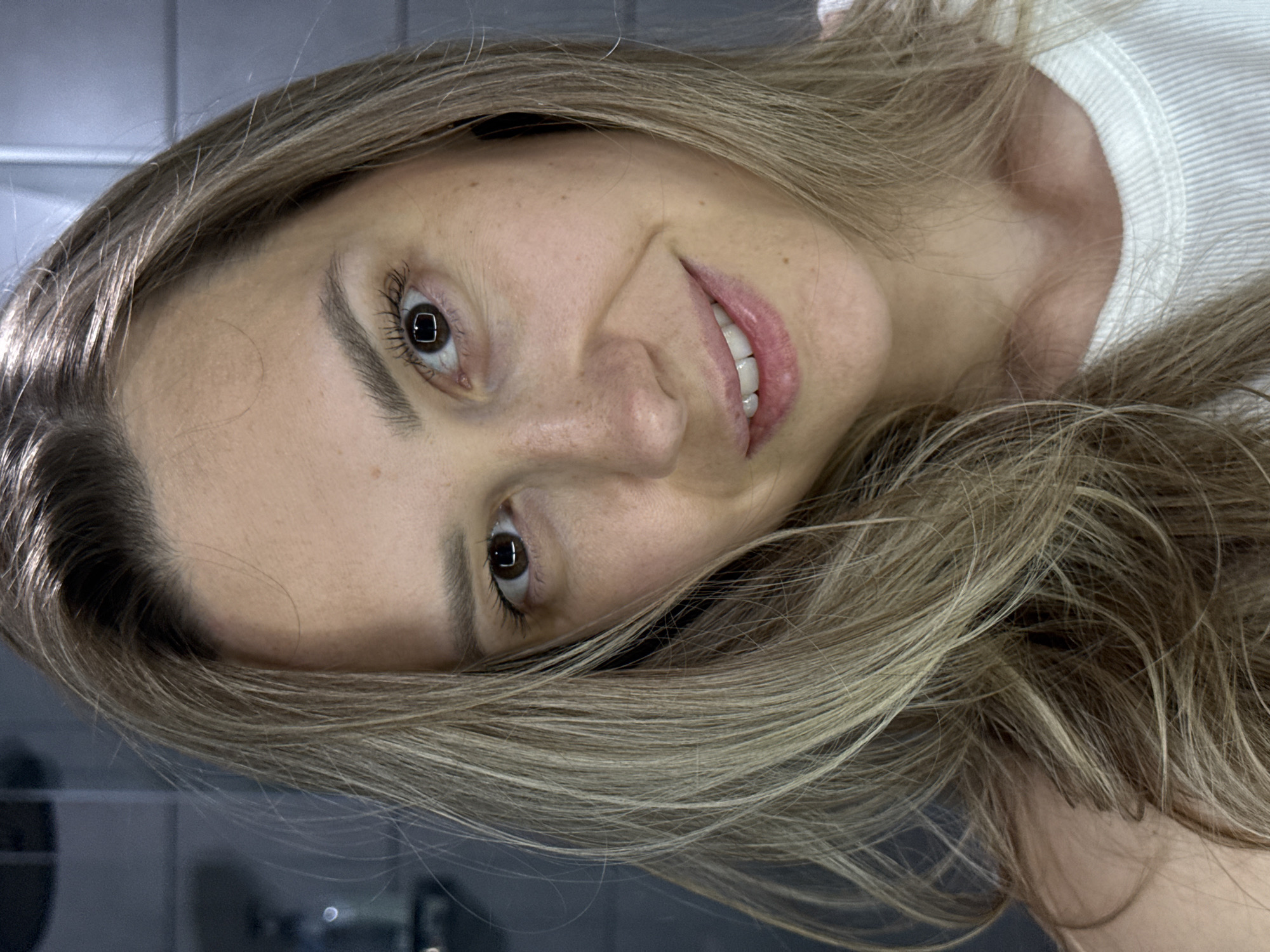 A close-up, tilted portrait shows a smiling woman with light brown hair. Her eyes are brown, and she has freckles on her face. She is wearing a white top. The background appears to be a gray wall, possibly in an indoor setting like a bathroom. The location in the city of Chaikovsky, Russia, cannot be determined from the image.