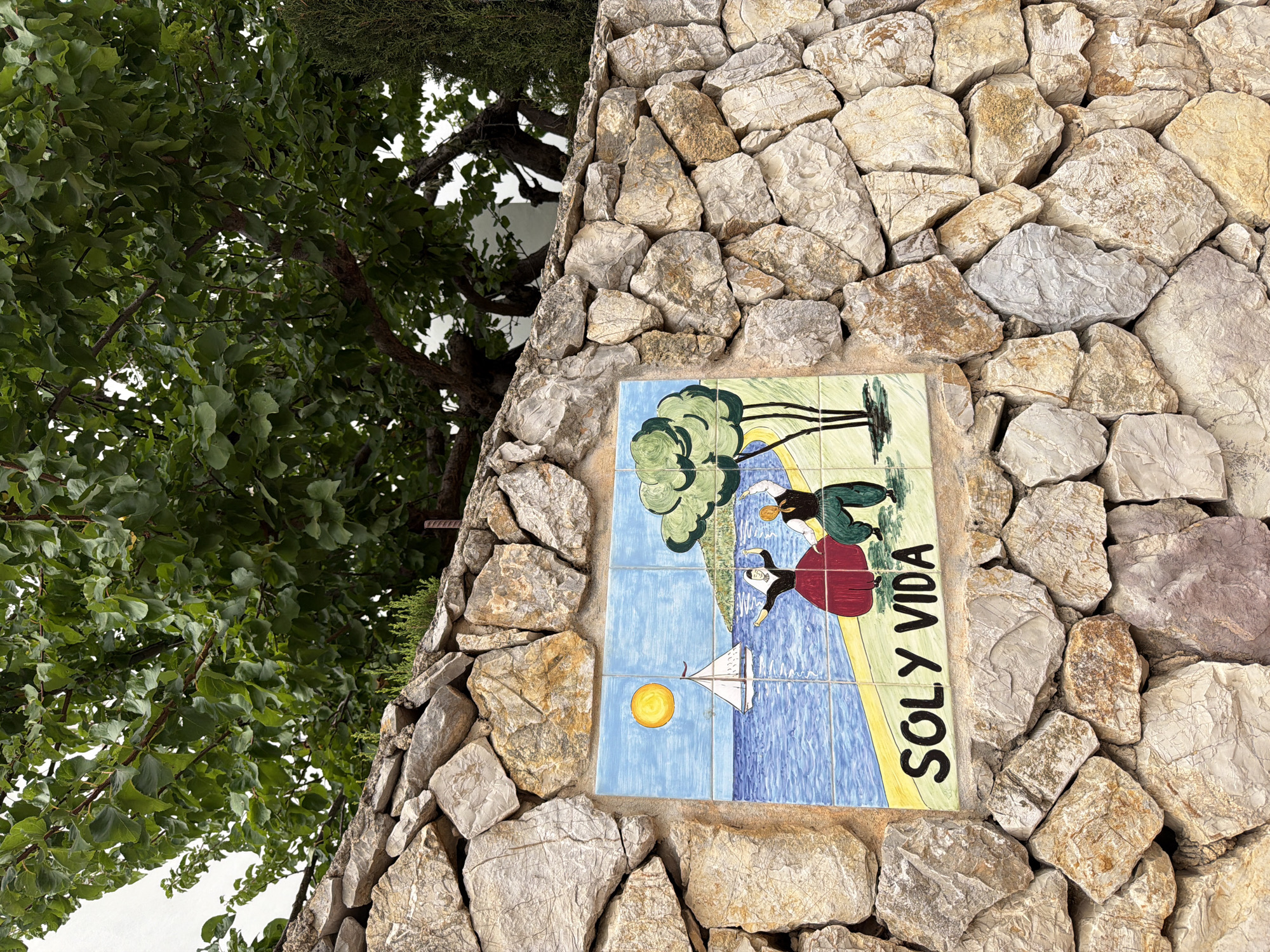 The image shows a ceramic tile mural attached to a rough stone wall. The mural depicts a seaside scene with a sailboat on blue water under a yellow sun. Two figures, one in a red dress and the other in green trousers and a black vest, are dancing on the shore next to a tree. The text "SOLY VIDA" is written in black letters below the figures. The stone wall is constructed from irregular, light-colored rocks. Lush green foliage from a tree hangs over the wall from above. The setting is outdoors, with a portion of a building visible in the background. This mural is located in Palmanova, Spain.