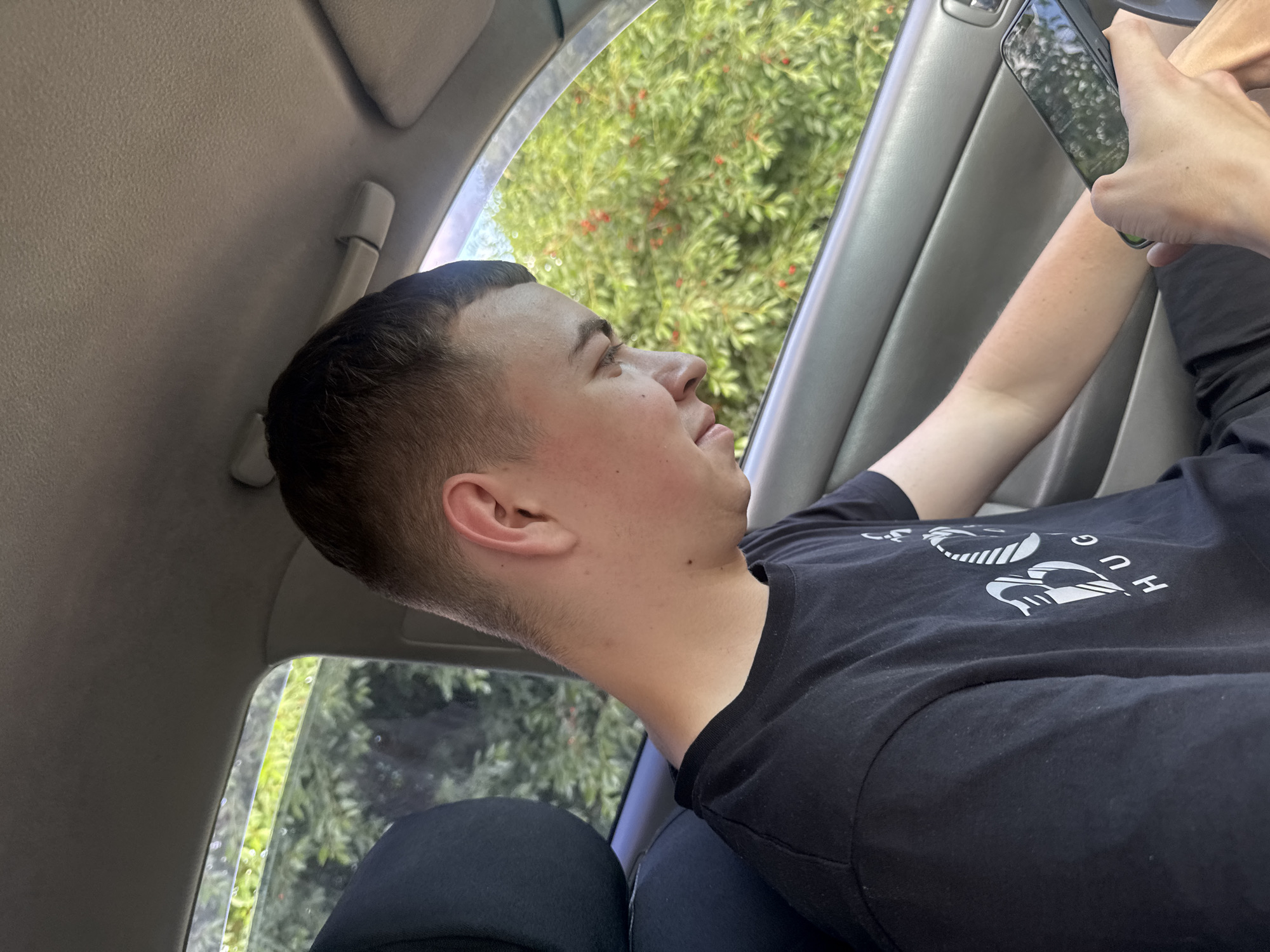 A young male with a buzz cut and wearing a black t-shirt with a white logo is seated in the passenger seat of a car, looking down at his phone. He is holding the phone in his right hand and his left arm rests on his lap. Outside the car window, green foliage with small red berries is visible. The interior of the car, including the door panel and part of the headrest, is grey. The scene appears to be in Bohunove, Ukraine.