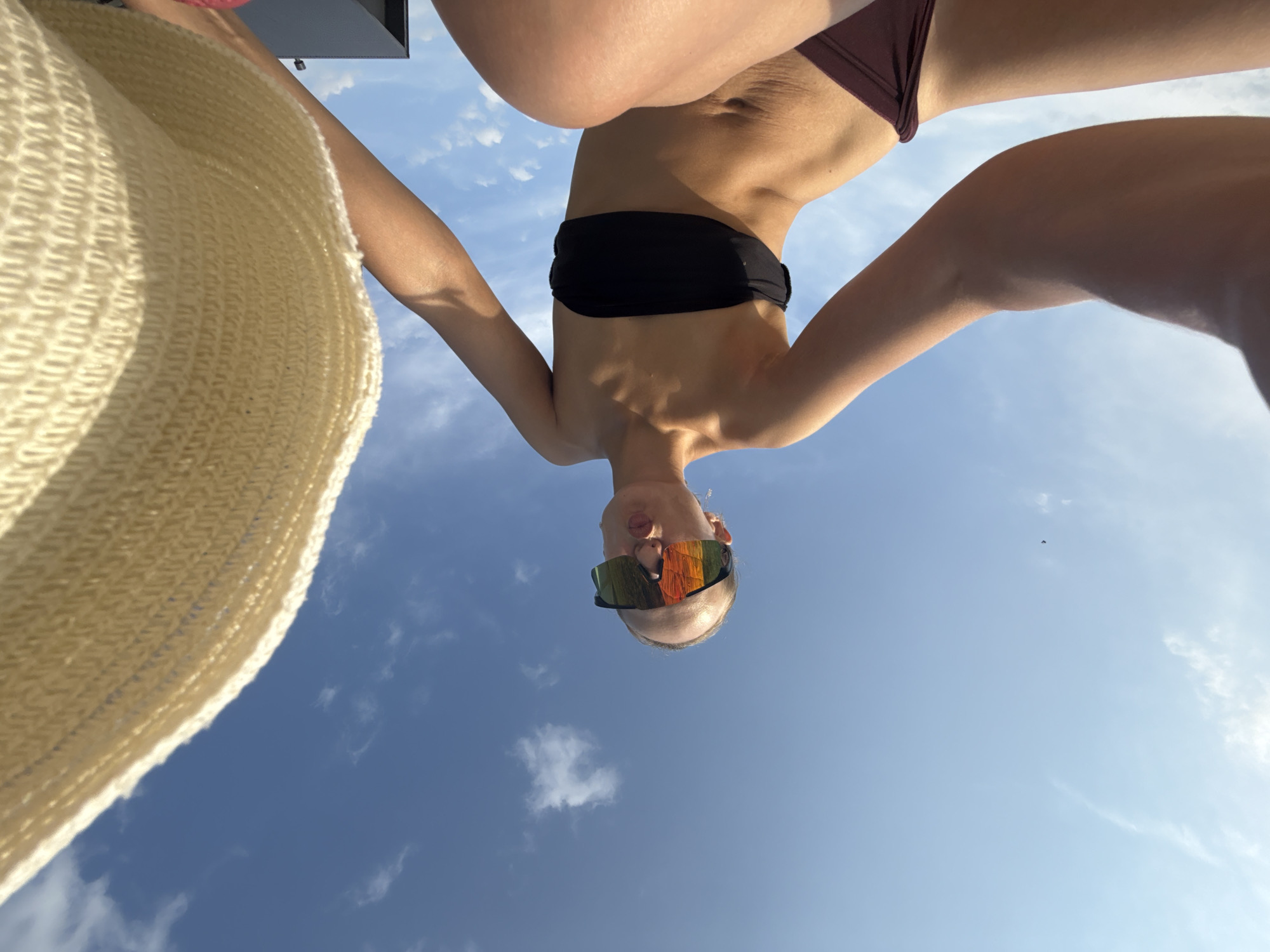 A low-angle, wide-shot selfie captures a woman in a bikini and large, reflective sunglasses posing against a bright blue sky with scattered clouds. The woman, with her hair pulled back, puckers her lips, looking directly at the camera. A portion of a wide-brimmed straw hat is visible in the foreground on the right side of the frame. The woman's arms are extended, with her left arm holding what appears to be the camera. The background is predominantly sky, with a small, dark object that could be a building visible in the lower right corner, along with a hint of pink fabric. The scene is outdoors, likely on a sunny day. There are no discernible landmarks or features that would place this image specifically in the city of Подшивалово, Russia.
