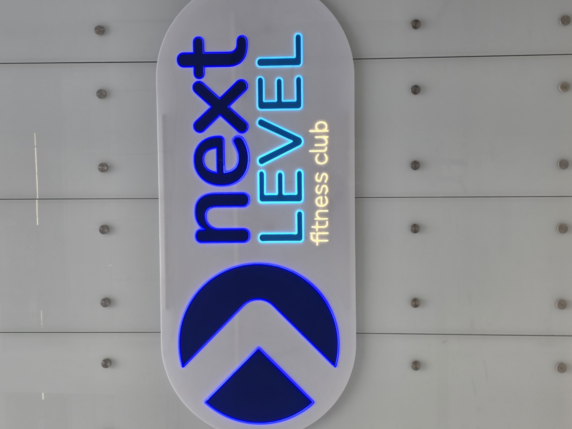 The image depicts a vertically oriented, rounded rectangular sign for "Next Level Fitness Club." The words "Next" are stacked vertically in blue neon lettering, followed by "Level" in cyan neon lettering. Below that, the words "fitness club" are rendered in a yellowish-gold, non-neon font. Beneath the text is a geometric logo comprised of a blue neon semicircle with an arrow shape inscribed within it, also in blue neon.
The sign appears to be mounted on a wall, which consists of horizontally oriented, frosted glass panels held in place by silver metal hardware. The location of this scene is in Plovdiv, Bulgaria.