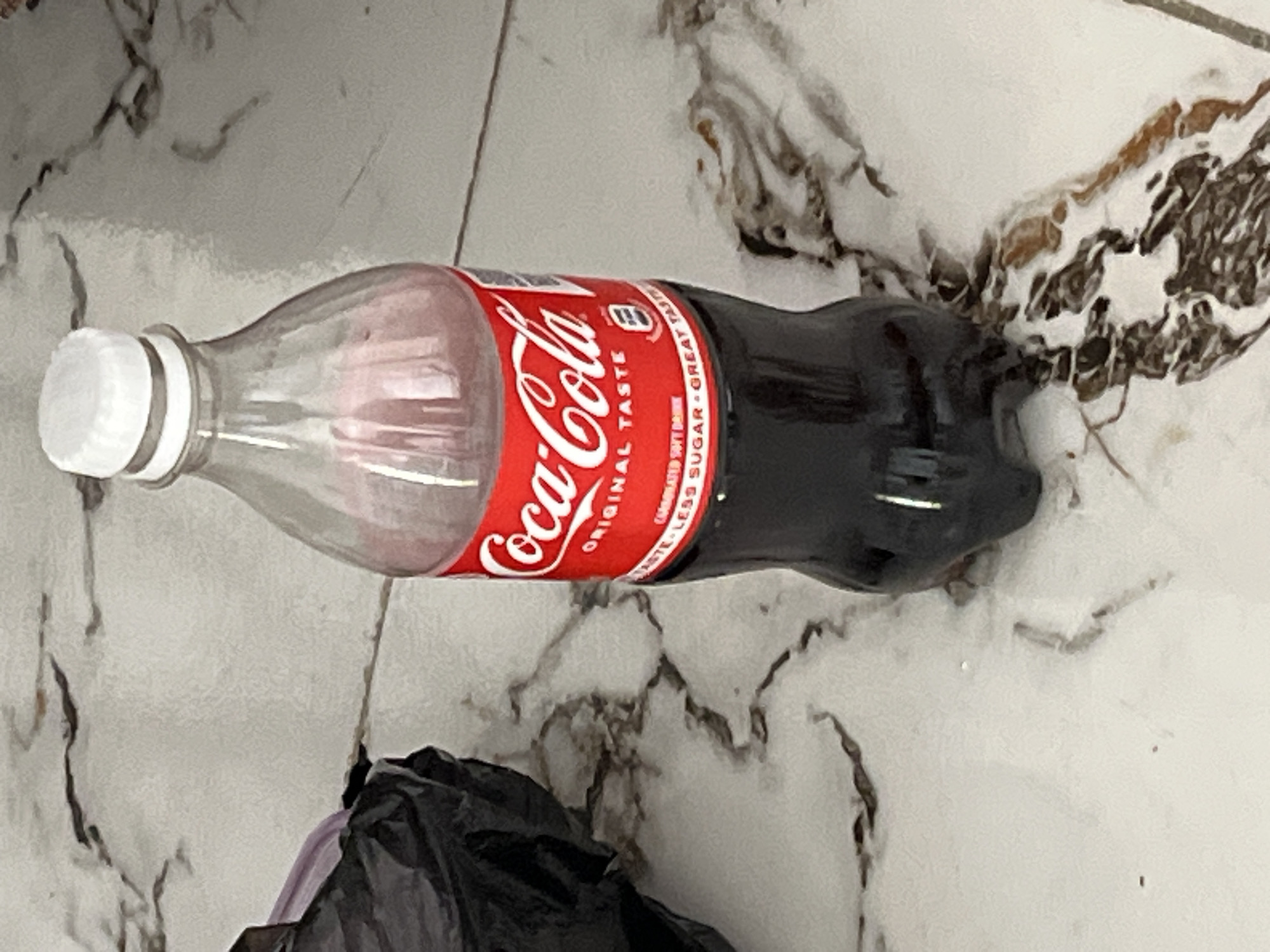 A partially-consumed bottle of Coca-Cola, labeled "Original Taste Great Taste Less Sugar," lies horizontally on a light-colored marble or tile floor. The bottle is plastic with a white cap and a red label featuring the Coca-Cola logo. It appears approximately half-full with a dark liquid. The scene is indoors. A portion of a black plastic bag is visible near the bottom of the image. The location is presumed to be in Jos, Nigeria.