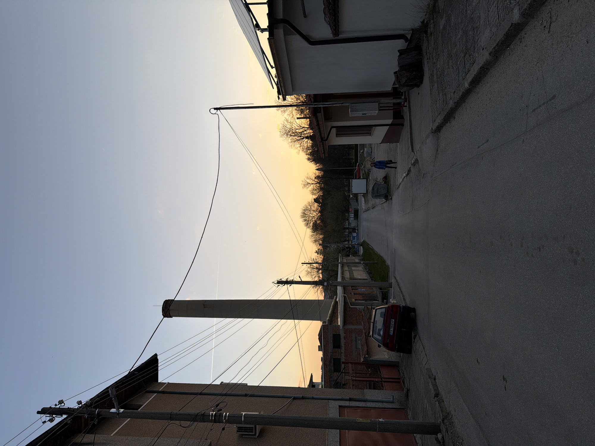 The image depicts a street scene in Panagyurishte, Bulgaria, during twilight. A tall, concrete chimney dominates the left side of the frame, towering over adjacent buildings and power lines. A dark red car is parked on the right side of the street. Two people, dressed in blue jackets, are visible in the distance, walking along the road. The road itself is paved and appears to be in an older residential area. Buildings on either side of the street vary in style, some being brick and others stucco. The sky is gradient, transitioning from light blue to a warm yellow-orange. Trees with bare branches stand in the middle ground along the road, likely within a park or green space.