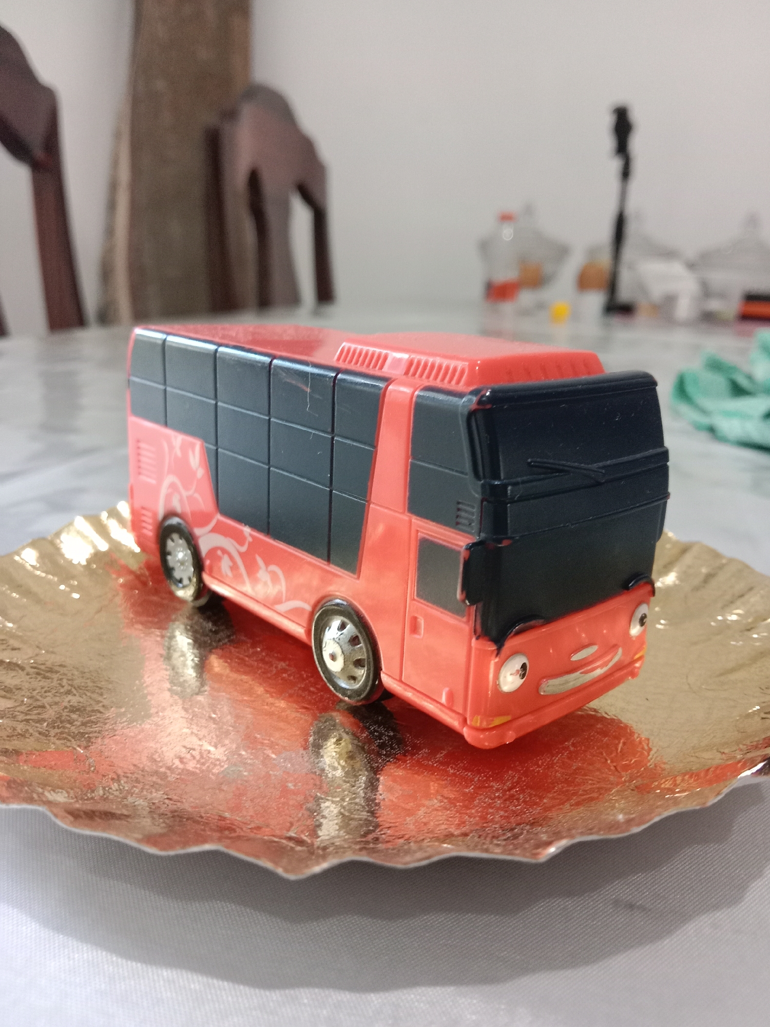 A close-up, eye-level shot shows a small, toy bus with a bright orange body and black windows. The bus has a cartoonish face with two large, round eyes and a smiling mouth. It is resting on a metallic gold, wavy-edged plate. The plate sits on a textured gray tablecloth. In the background, out of focus, are dark wooden chairs and a blurry white surface with some indistinct objects. The lighting is soft, coming from the front, and casts a reflection of the bus onto the plate. The overall impression is that of a still life arrangement, possibly on a dining table, in an indoor setting. There is no indication of the location being in Ngingas, Indonesia, or any other specific city.