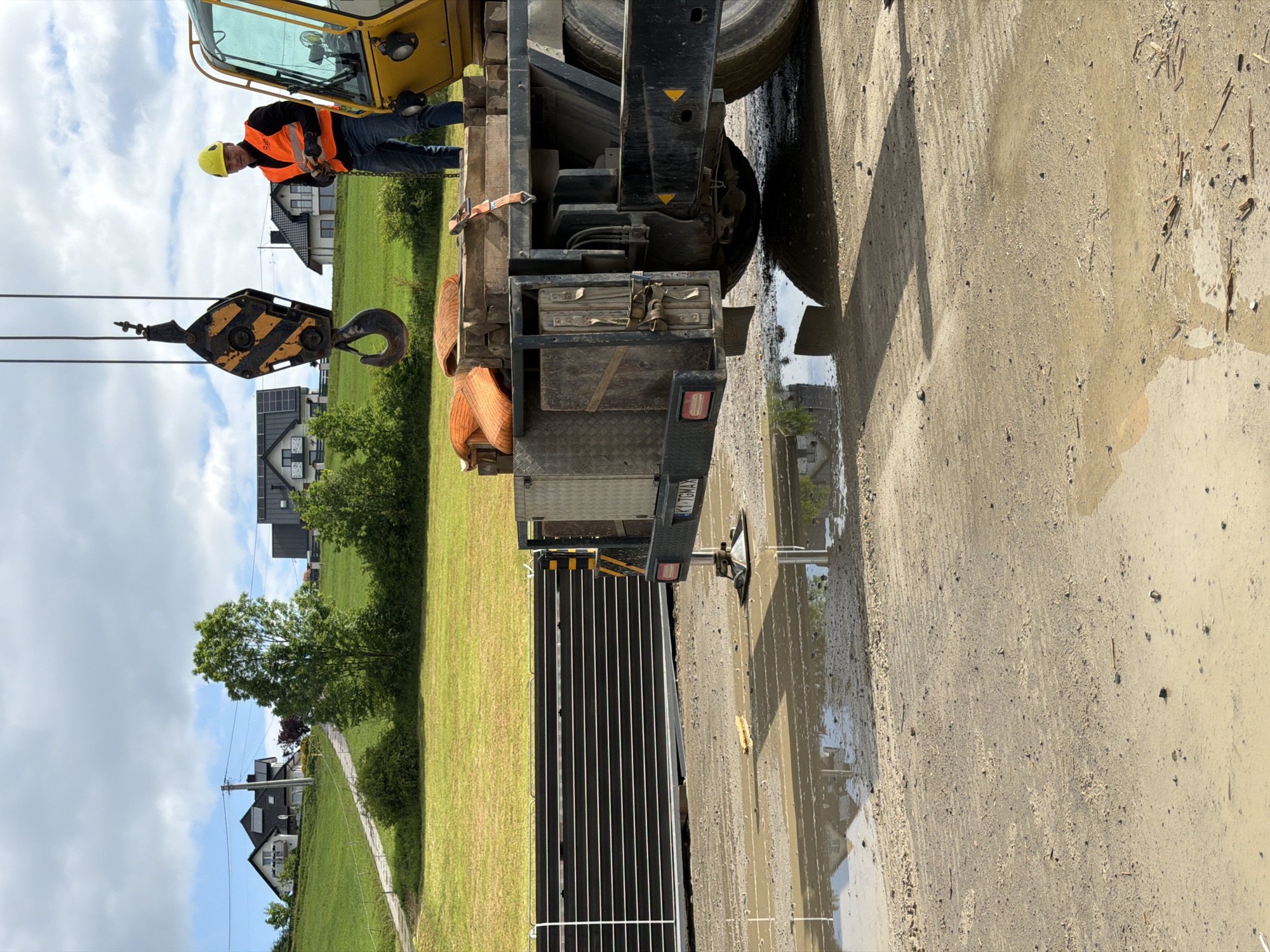 The image depicts a construction site in Męcina, Poland. In the foreground, a muddy ground with puddles is visible. To the left, a long, black, rectangular structure is partially submerged in mud. In the center and right, a yellow construction vehicle, likely a crane, is present. A worker wearing a yellow helmet and an orange safety vest stands on the platform of the vehicle, holding something in their hands. A large hook and pulley system hangs from the crane. In the background, a grassy hill with houses and trees is visible under a partly cloudy sky. The overall scene suggests ongoing construction or material unloading activity.