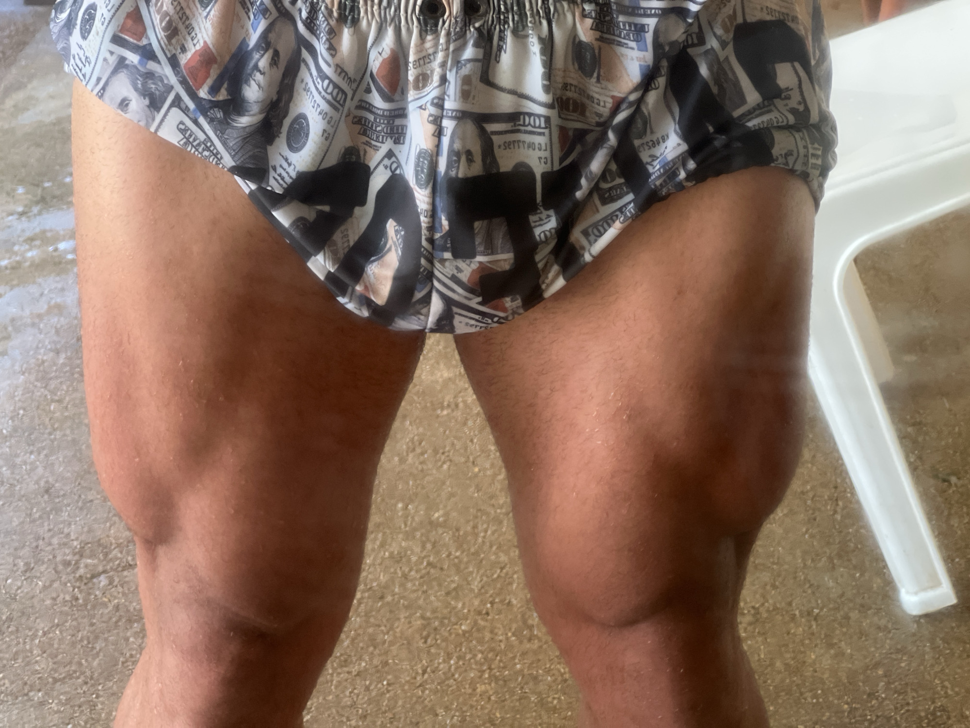 A close-up, low-angle shot shows the lower torso and upper legs of a person standing on a tan, textured surface. The person is wearing patterned boxer briefs featuring an image of US hundred-dollar bills and black abstract shapes. The person's skin is tanned and appears moist. To the right of the frame, a portion of a white plastic chair is visible. The background is out of focus, but the ground appears to be a paved or tiled area.
