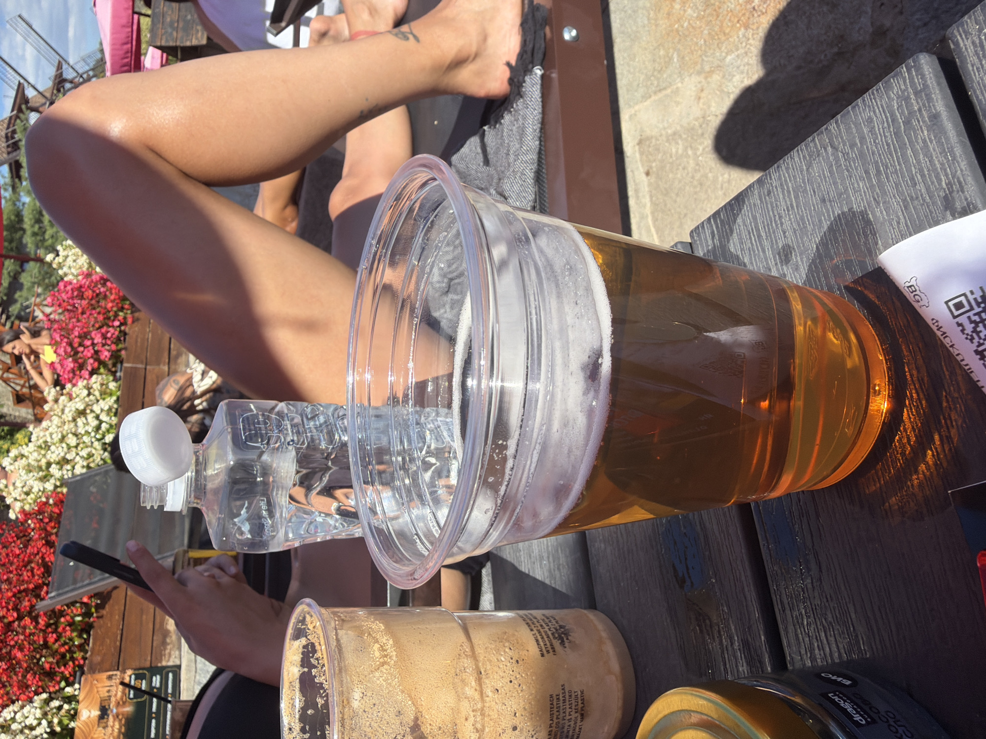 In Gorna Malina, Bulgaria, on a wooden table outdoors, a plastic cup filled with amber liquid and white foam sits in the foreground. To the left, another plastic cup contains a frothy brown beverage. Behind these, a clear plastic bottle of water with an open white cap is visible. In the background, a person's legs are extended, with floral decorations and outdoor seating suggesting a resort or recreational area. A hand holding a smartphone is also visible. The lighting indicates a sunny day.