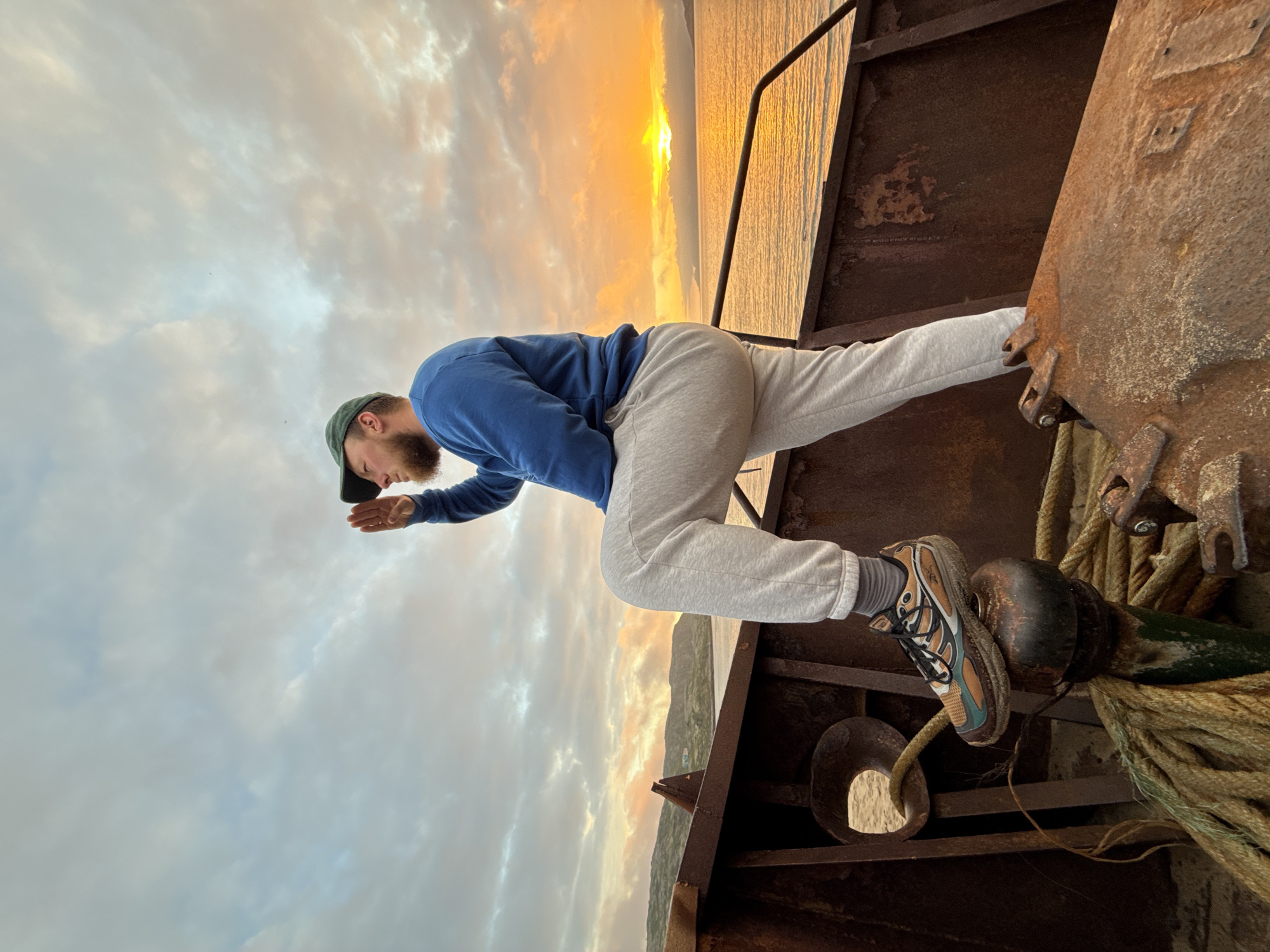 A man wearing a blue sweatshirt, grey sweatpants, a green baseball cap, and athletic shoes stands on the rusty deck of a shipwreck. He is looking out at the ocean, with his right hand raised to shield his eyes from the setting sun. The sun is low on the horizon, casting a warm glow over the water and the surrounding landscape. The man is standing with his left leg bent at the knee, with his foot resting on a metal capstan. A thick rope is coiled around the capstan. The shipwreck appears to be beached on a sandy shore, with hills in the background. The sky is filled with clouds, some of which are illuminated by the setting sun. The scene is set in Teriberka, Russia, a coastal village known for its shipwrecks and dramatic natural beauty.