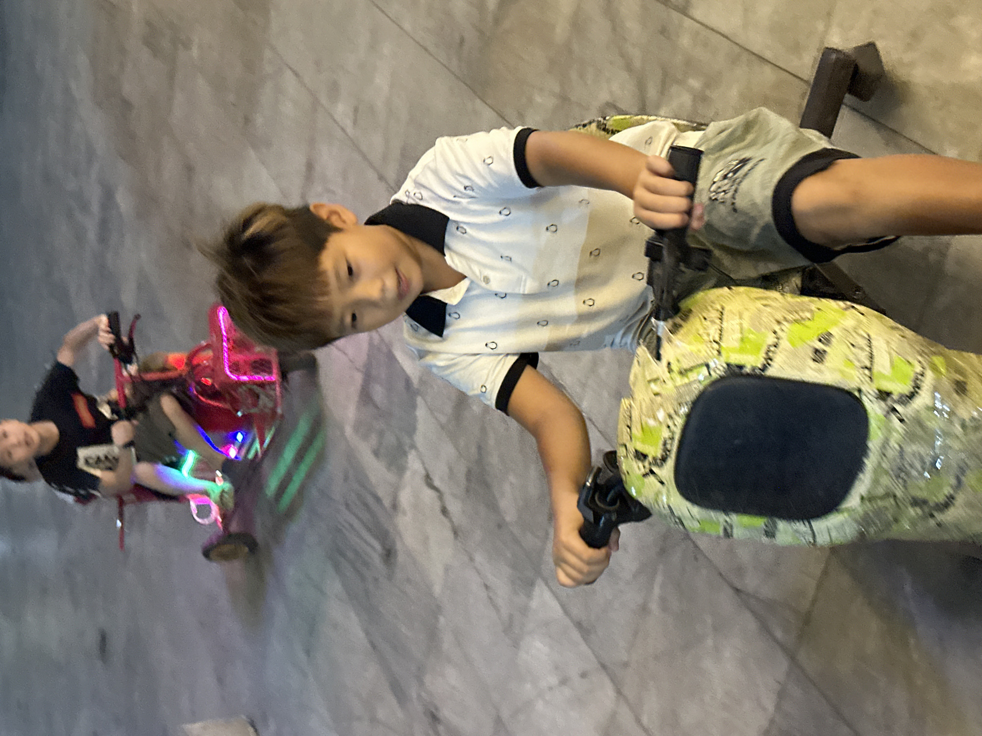 This tilted image, captured in Phố Ràng, Vietnam, features two boys riding electric drift trikes on a light grey, possibly tiled or concrete surface, against a plain grey wall background.
In the foreground, a boy with short brown hair is smiling and looking towards the camera. He is dressed in a white polo shirt with black trim and small circular patterns, paired with light-colored shorts. He is actively steering his drift trike, which has a distinct green and yellow camouflage-like pattern on its body and a black seat.
Further back and to the left, a second boy is also engaged in riding a drift trike. He is wearing a dark t-shirt with an unidentifiable white graphic or text, and dark shorts. His trike stands out with its bright red/pink components and vibrant multi-colored LED lights (blue and green), suggesting a recreational setting that might be indoors or during evening hours. Both boys appear to be enjoying the activity. No clearly legible text is visible on clothing or equipment.