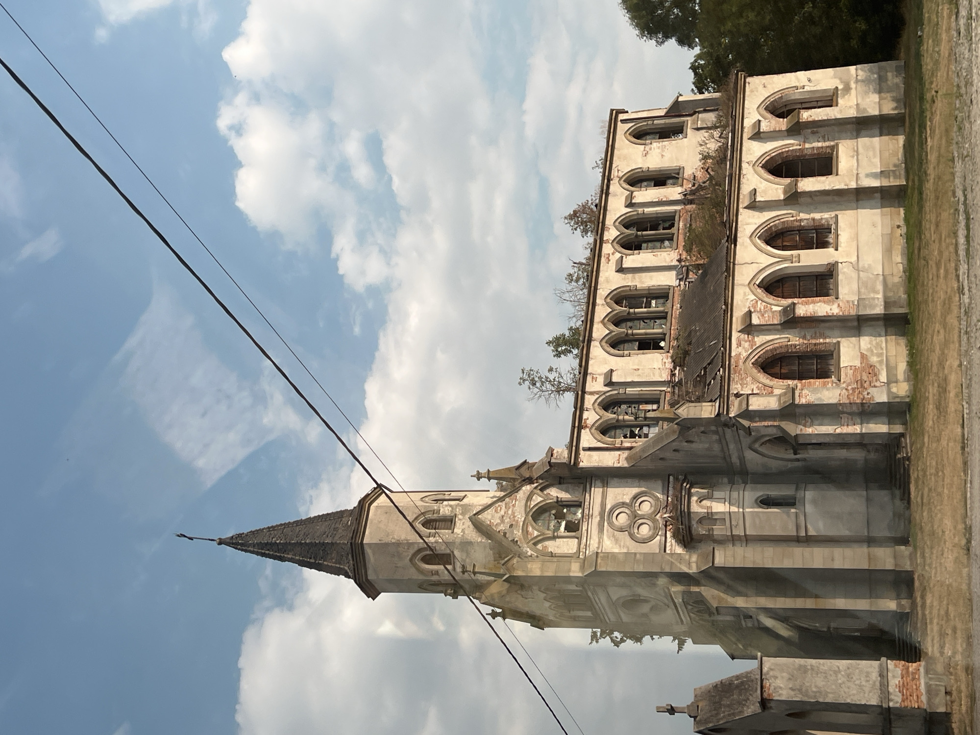 In the city of Sorotske, Ukraine, an abandoned church stands under a sky with scattered clouds. The church is a Gothic-style structure with a tall, pointed spire topped with a cross. The building is in a state of disrepair, with broken windows, exposed brickwork, and vegetation growing from the roof. The exterior walls are made of light-colored stone, weathered and cracked in many places. In the foreground, dry grass covers the ground. A power line stretches diagonally across the upper portion of the image, obscuring a portion of the sky and the church's spire. To the left of the main structure, a smaller, detached building resembling a chapel or mausoleum with a cross on top is visible. The scene suggests a forgotten and neglected place, weathered by time and the elements.