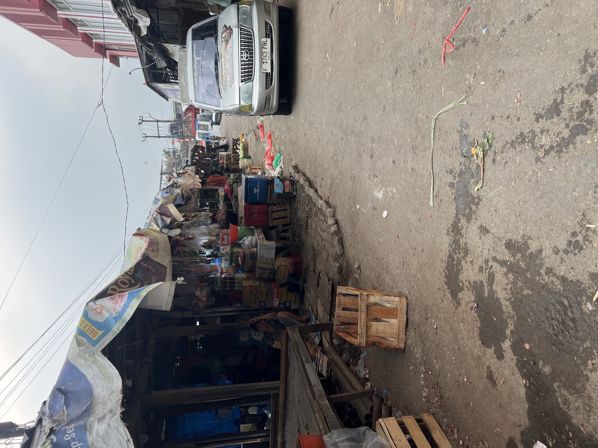The image depicts a street market scene in Citeureup, Indonesia, under an overcast sky. A silver Toyota Kijang van, with license plate F 1745 FNL, is parked on the right side of a dirt and asphalt road. The van has decorative stickers on its hood. To the left of the van, a row of market stalls with makeshift roofs made of corrugated metal and tarps lines the street. These stalls are filled with various goods, including what appears to be produce, packaged items, and some plastic bags.
In the foreground, a wooden crate is positioned on the ground near the stalls. The ground is uneven, with patches of wetness and scattered debris, including plastic bags and what look like onion skins. The market appears to be set up along a busy road, with overhead power lines crisscrossing the sky. In the background, buildings with red and white facades are visible, suggesting the market is situated within an urban or semi-urban area. Several people are present, some appearing to be vendors or shoppers, though their interactions are not clearly discernible. The overall impression is one of a bustling, open-air market operating in a somewhat informal setting.