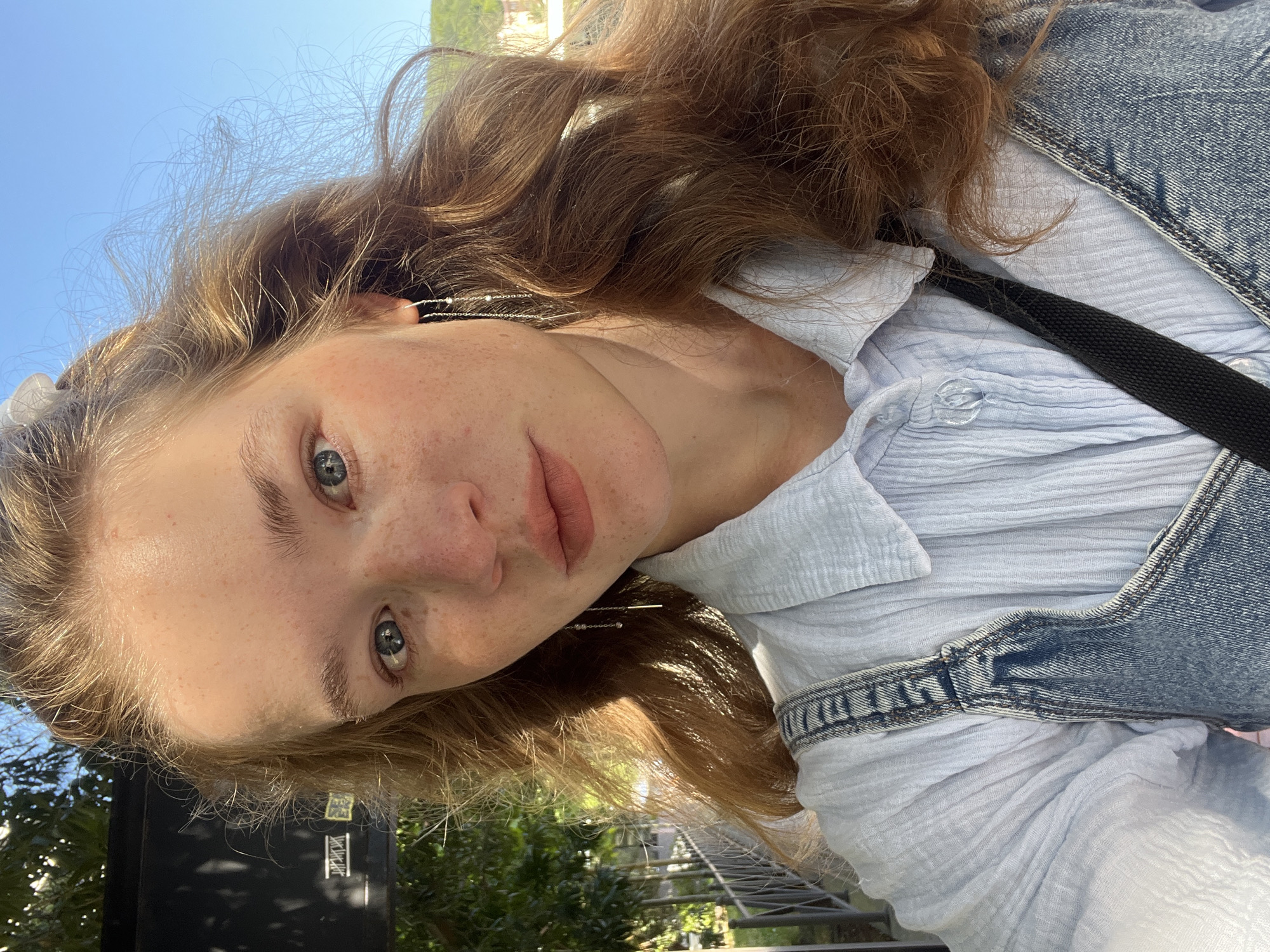 A young woman with light skin, freckles, and light brown hair is photographed at an angle. She has blue eyes and light pink lips. She is wearing a light blue, textured, button-down shirt with denim overalls. A black strap runs across her chest. She is outdoors in daylight, with a blue sky visible in the background. There are trees and foliage visible in the lower portion of the image. The location is likely Budva, Montenegro, as indicated in the prompt, though specific location details are not evident in the image.