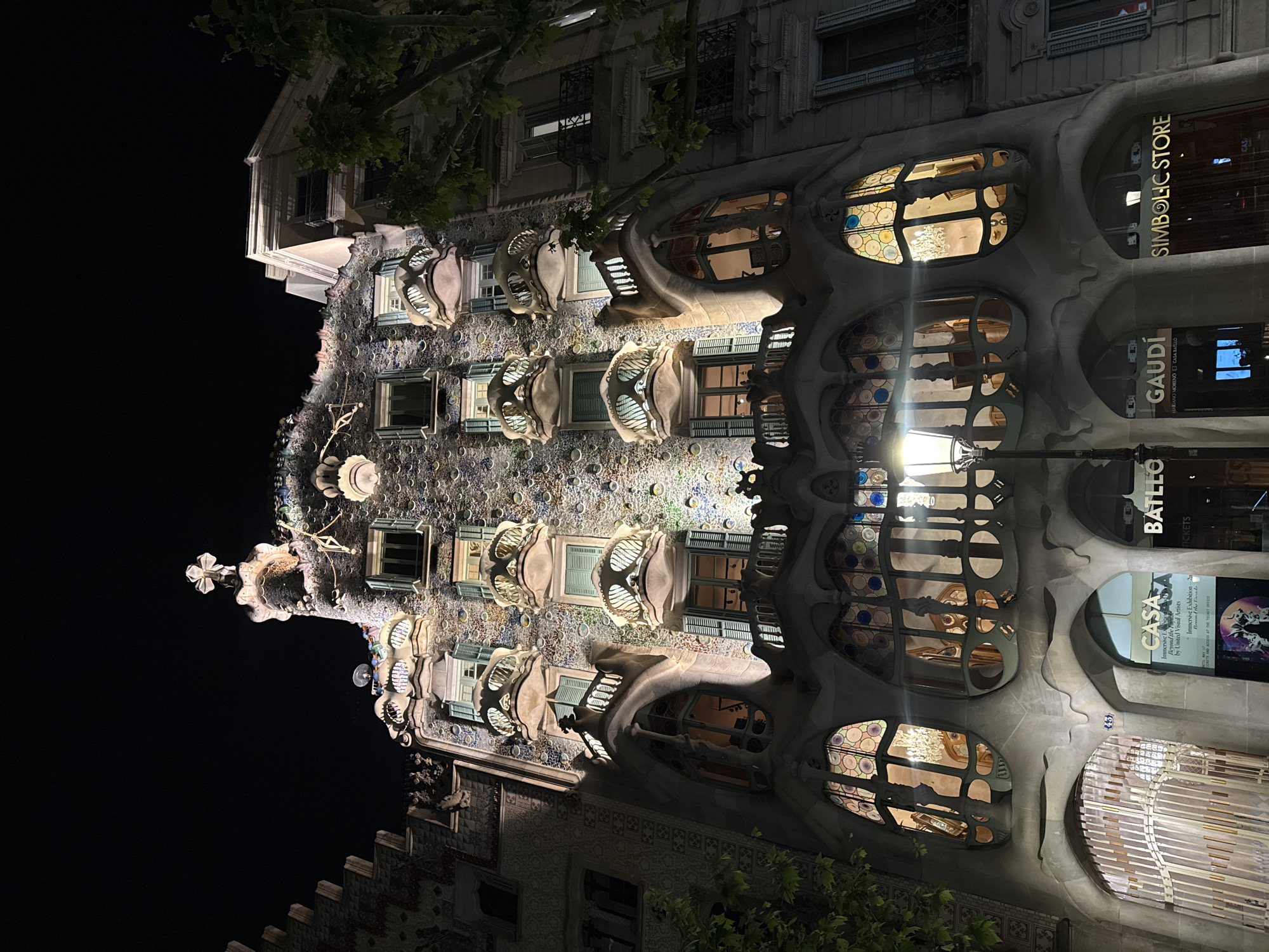 The image showcases Casa Batlló, a renowned building designed by Antoni Gaudí, located in Barcelona, Spain. It's captured at night, with the building illuminated. The distinctive façade, with its skeletal balconies and colorful mosaic patterns, is the focal point. Signs advertising "Casa Batlló Tickets" and "Symbolic Store" are visible, suggesting it is a tourist attraction. A lamp post stands in front of the building, providing further illumination.
