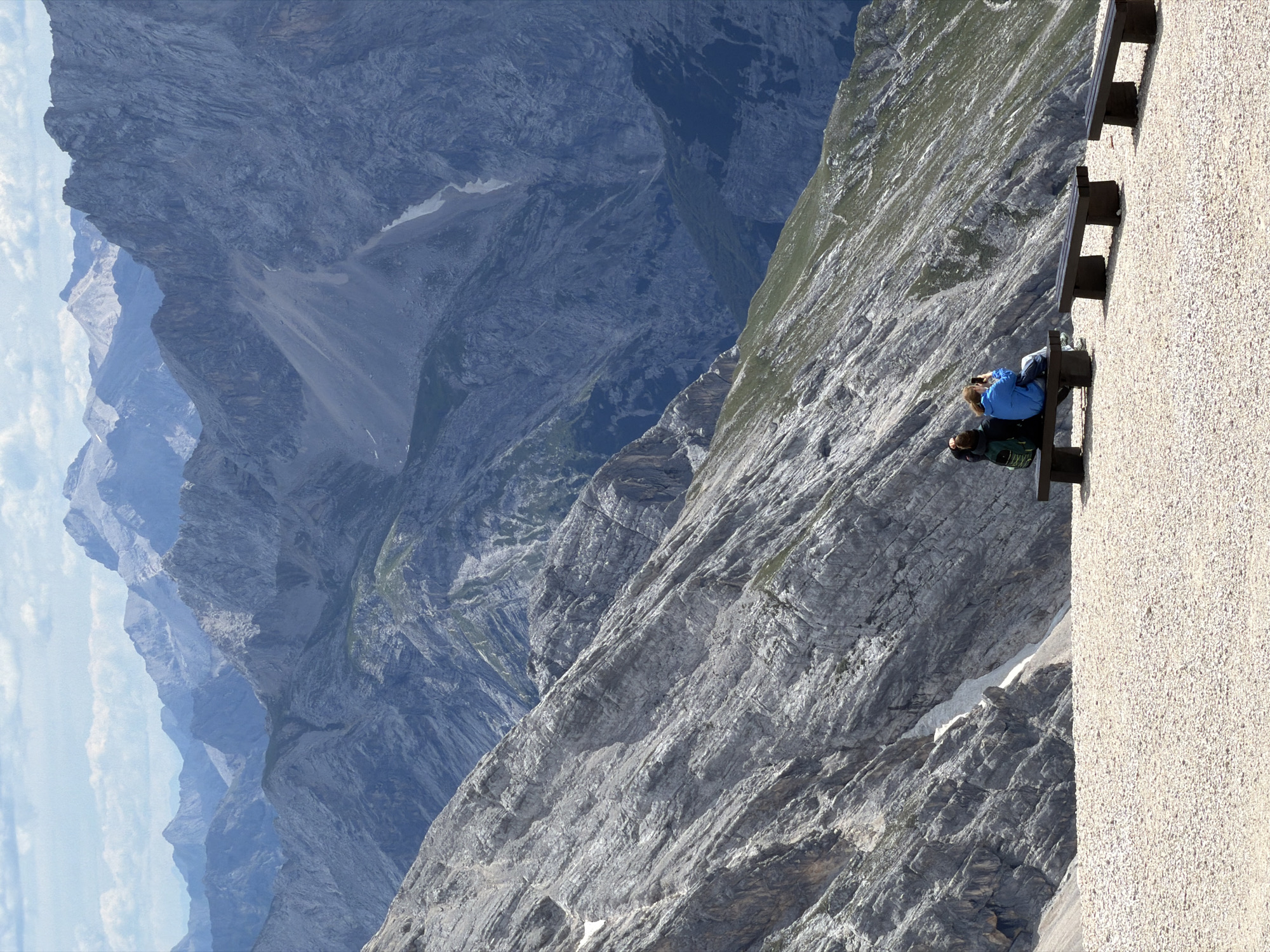 The image depicts a scenic mountain landscape, likely the Alps, with rugged, grey rock formations and patches of green vegetation. In the foreground, a gravelly area is furnished with three dark wooden benches. Two individuals are seated on the leftmost bench. One person, wearing a blue jacket and jeans, is holding a phone and appears to be taking a photo or video. The other person, with a backpack, is also seated, facing away from the camera. The background is dominated by layers of majestic, blue-hued mountain ranges under a cloudy sky. This scene is representative of viewpoints found in the vicinity of Garmisch-Partenkirchen, Germany, known for its access to alpine vistas.