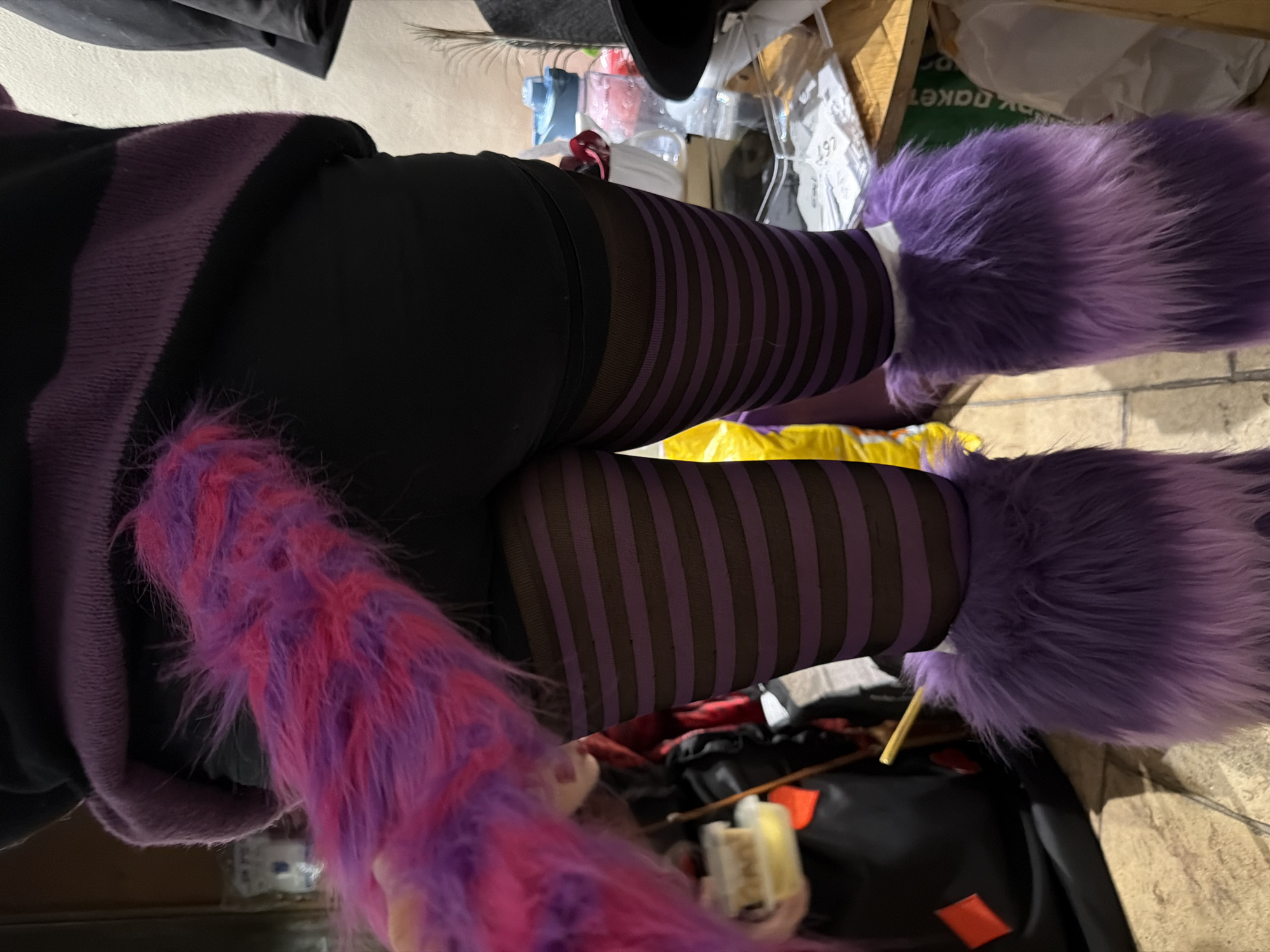 An indoor, eye-level shot captures a person from the waist down, likely showcasing a costume. The subject wears black pants or leggings, covered from the knees downwards by striking purple and black striped tights or stockings. Over these, fluffy purple leg warmers or boot covers are worn, extending from the feet upwards. A long, shaggy tail, predominantly pink with streaks of purple, hangs down on the left side of the frame. Part of a dark purple hooded garment, possibly a sweatshirt or jacket, is visible around the waist area.
The background is somewhat cluttered, revealing various objects. On the upper right, there appears to be a black hat, a feather, and numerous items on shelves or a surface, including plastic containers, bags, and boxes. One white package or bag in the background has partially legible text, including what looks like "dakeT" (when rotated) and the numbers "197". On the floor or a lower surface, there are more items, including a yellow bag, a black container with orange accents, and a wooden stick. The lighting suggests an indoor setting, possibly during the day or under artificial light. No specific activity beyond posing is evident, and emotions are not discernible. The location context is Nir Eliyahu, Israel.