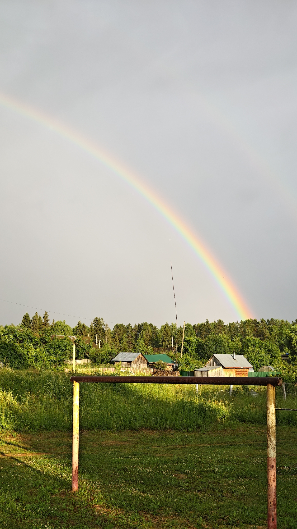 This image depicts a rural landscape in Уральское, Russia, under a partly cloudy sky where a vibrant rainbow arcs across the horizon. The foreground is dominated by a grassy field with a rustic metal structure, possibly part of a goalpost or fence, with two weathered poles supporting a horizontal bar. The grass is lush and green, dotted with small white flowers, and bathed in sunlight that casts long shadows.
In the mid-ground, a dense line of green trees forms a natural backdrop, with hints of a small village nestled among them. Several wooden houses with different colored roofs are visible, suggesting a tranquil, lived-in setting. A tall, thin pole, possibly an antenna or part of a utility line, rises vertically from the tree line, breaking the otherwise natural horizon. A few tiny black specks in the sky, likely birds, are also present.
The rainbow is the most striking feature, its colors bright and distinct against the muted gray sky. This suggests that it has recently rained, but the sun is now breaking through, a common weather phenomenon in many regions. The time of day appears to be late afternoon or early evening, given the angle of the sunlight and the length of the shadows. The overall mood is serene and picturesque, evoking a sense of natural beauty and the quiet charm of rural life. There is no visible text in the image.