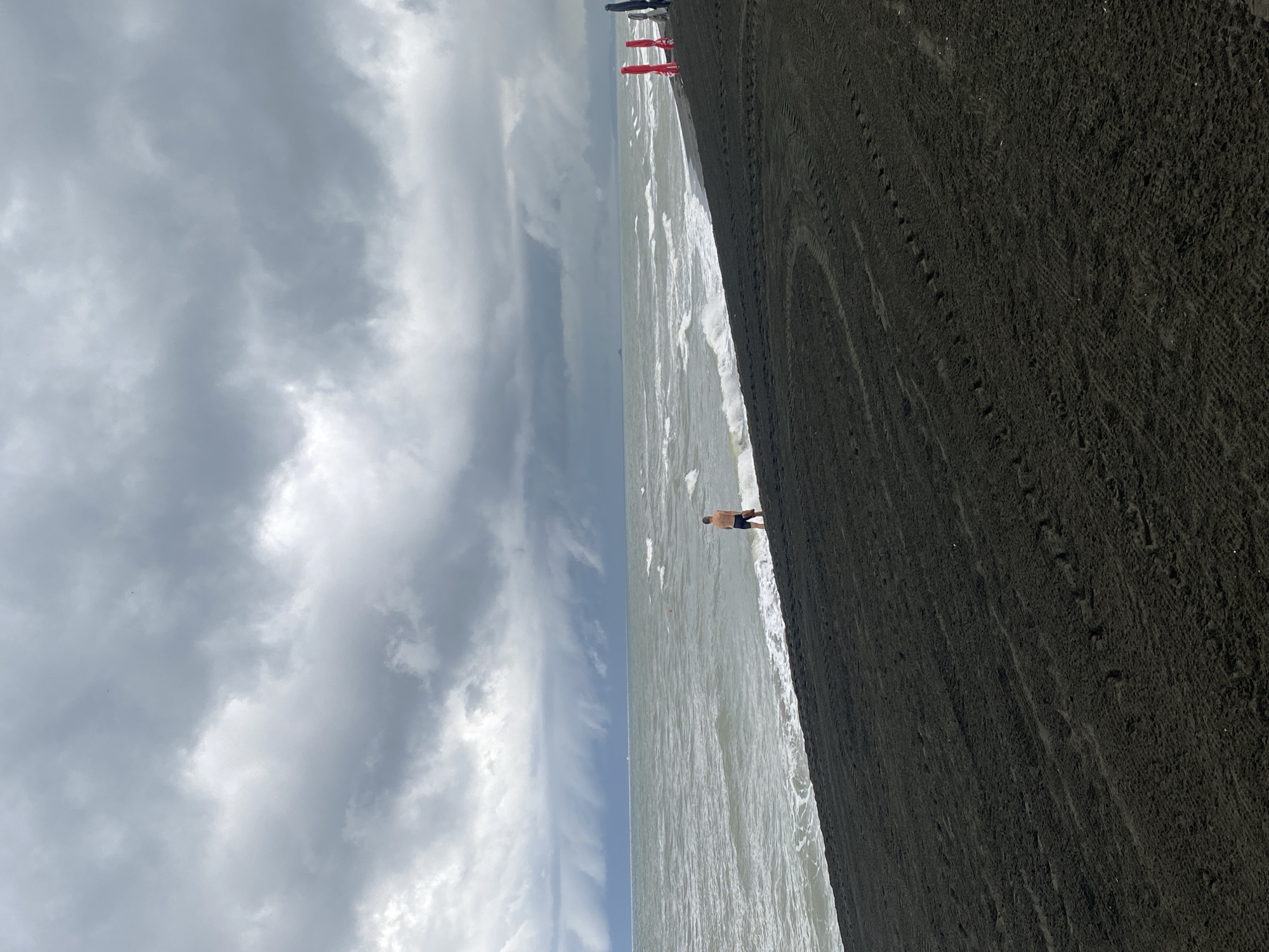 The image depicts a beach scene in Ureki, Georgia, characterized by dark sand and choppy, gray-green water under a heavily overcast sky. A lone man, wearing dark shorts, stands at the edge of the water, facing away from the camera. The waves are breaking on the shore, creating a foamy white line. Tire tracks are visible in the dark sand of the foreground, indicating recent vehicle activity on the beach. In the distance, a ship can be seen on the horizon, and several red and black umbrella-like structures are present on the beach to the right of the frame. The overall mood of the image is somber, likely due to the cloudy weather.