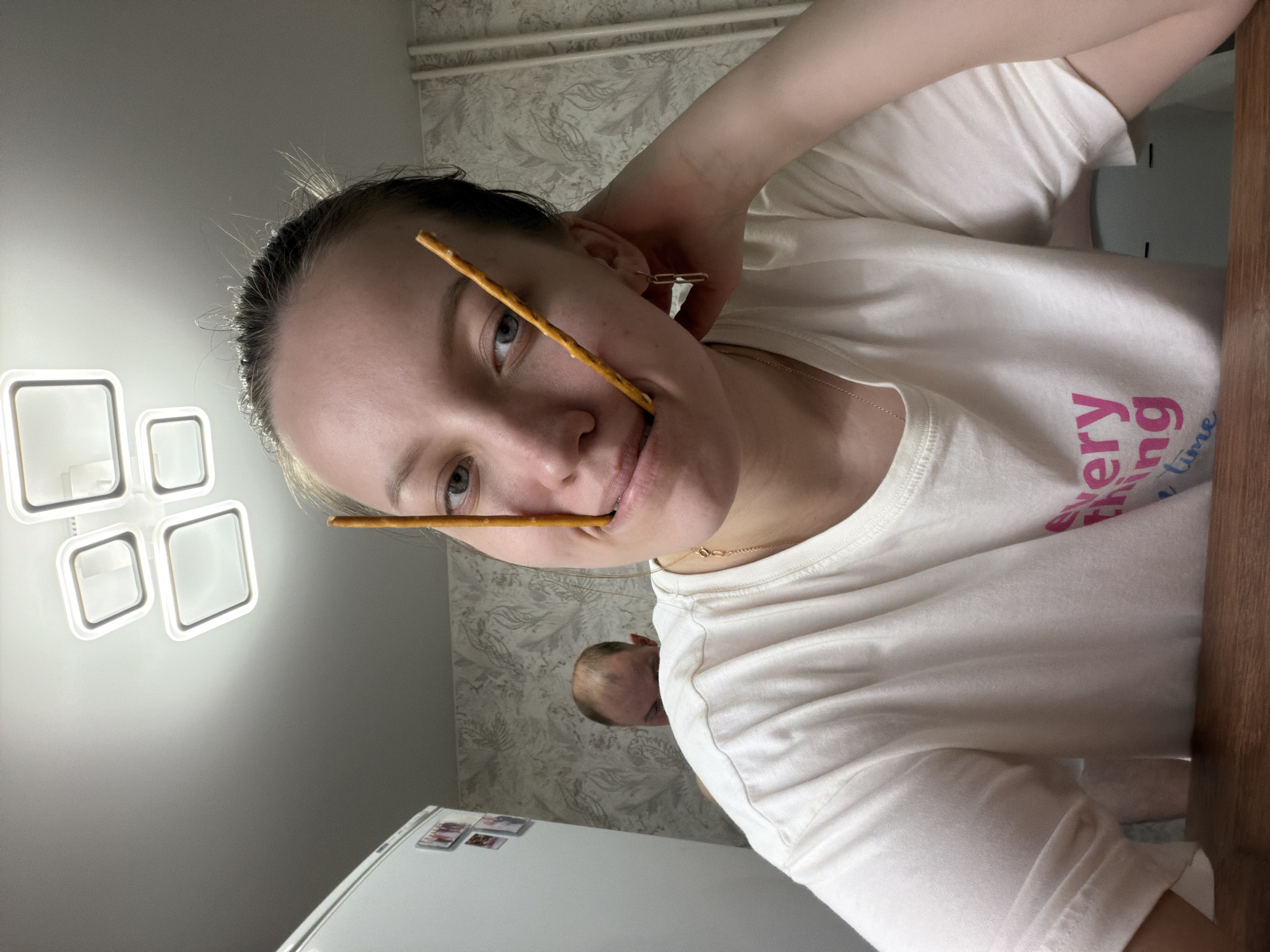 A person with light skin and hair pulled back is facing the camera, holding two breadsticks in their mouth. The background includes a light fixture, patterned wallpaper, a refrigerator, and another person in the background. It is impossible to confirm this photo was taken in Reshetnikovo, Russia.