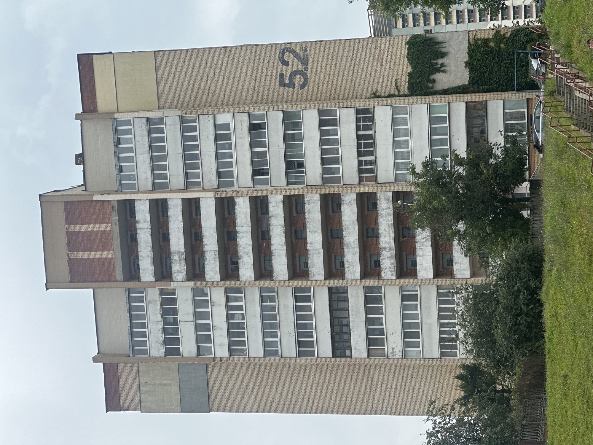 A tall, multi-story apartment building stands against a cloudy sky in Minsk, Belarus. The building's facade is a combination of light-colored brick and grey concrete panels. Numerous balconies with white railings run horizontally across the building. The number "5.2" is painted in dark grey on the brick section of the building. Green ivy covers a portion of the facade on the right side. Trees and green grass are visible in the foreground, with a wooden fence and a set of outdoor stairs leading up to the building. Cars are parked at the base of the building.
