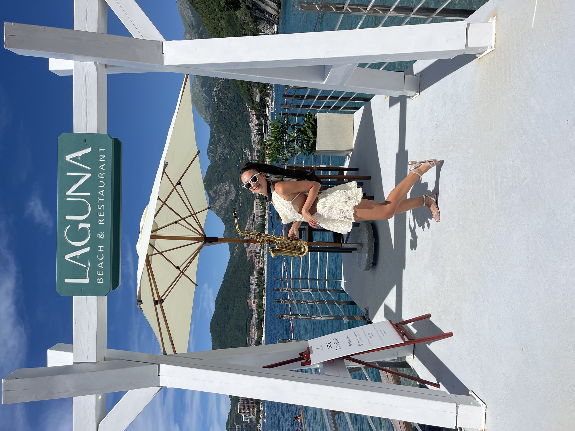 In Budva, Montenegro, a woman stands on a white deck, holding a saxophone. She wears a white lace dress and white sunglasses. Behind her, a sign reads "LAGUNA BEACH & RESTAURANT." The turquoise water of the Adriatic Sea stretches out before her, with mountains in the background. A white umbrella provides shade.