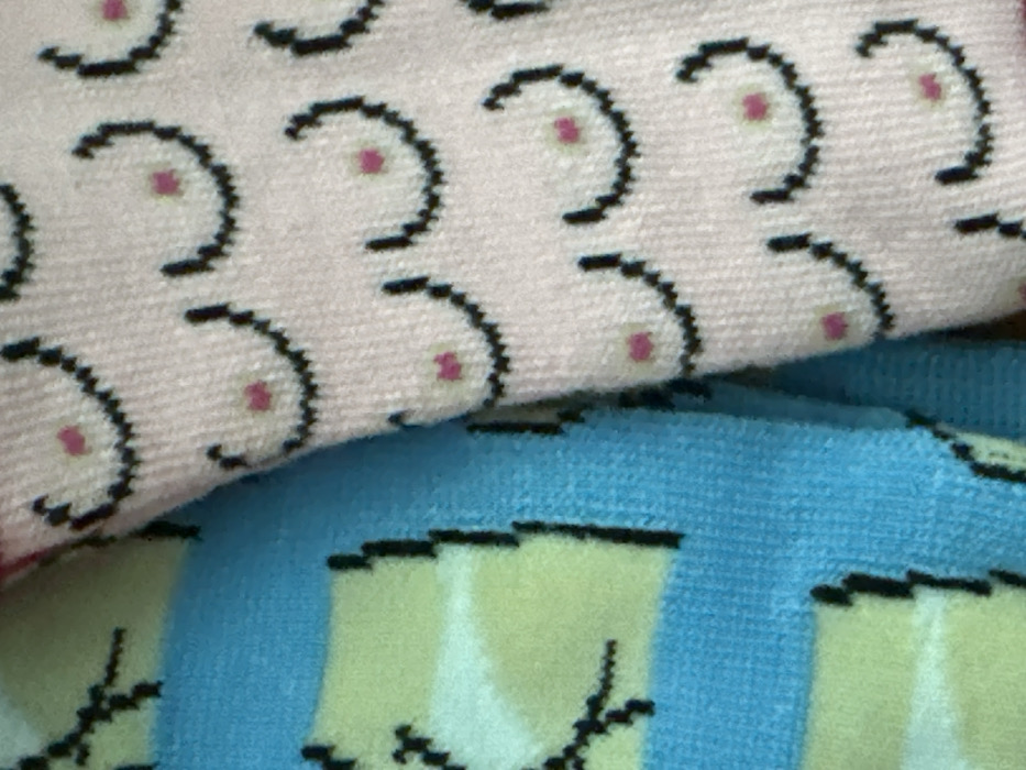 This is a close-up, slightly angled shot of two distinct pieces of patterned fabric, likely socks or similar textile items, layered or folded.
The top fabric, which occupies the majority of the upper half of the image, is a light, muted pink or off-white color. It features a repeating pattern of stylized black crescent shapes, resembling the letter 'C' or a half-circle, with a small, solid pink circular dot positioned just inside the open curve of each crescent. These motifs are arranged in an offset grid, creating a dense pattern across the fabric. The texture of this fabric shows a visible weave, suggesting it is a knitted material like cotton or a synthetic blend often used for socks.
Beneath and partially obscured by the top fabric is a second piece of textile. This bottom fabric is a vibrant light blue. It also displays a repeating pattern, which consists of light yellow or pale green rectangular or irregular shapes outlined in black. Some black lines extend from these shapes, appearing like slender, abstract appendages or decorative elements. The overall pattern on the blue fabric is less distinct than the top one due to cropping and slight blur, but it clearly uses a contrasting color scheme against the blue background.
Both fabrics appear soft and textured. No specific activity or event is taking place, as the image focuses solely on the fabric patterns. There are no people, explicit location details, or visible text in the image. The lighting is soft and even, without strong shadows, indicating it could be indoor lighting or an overcast day.