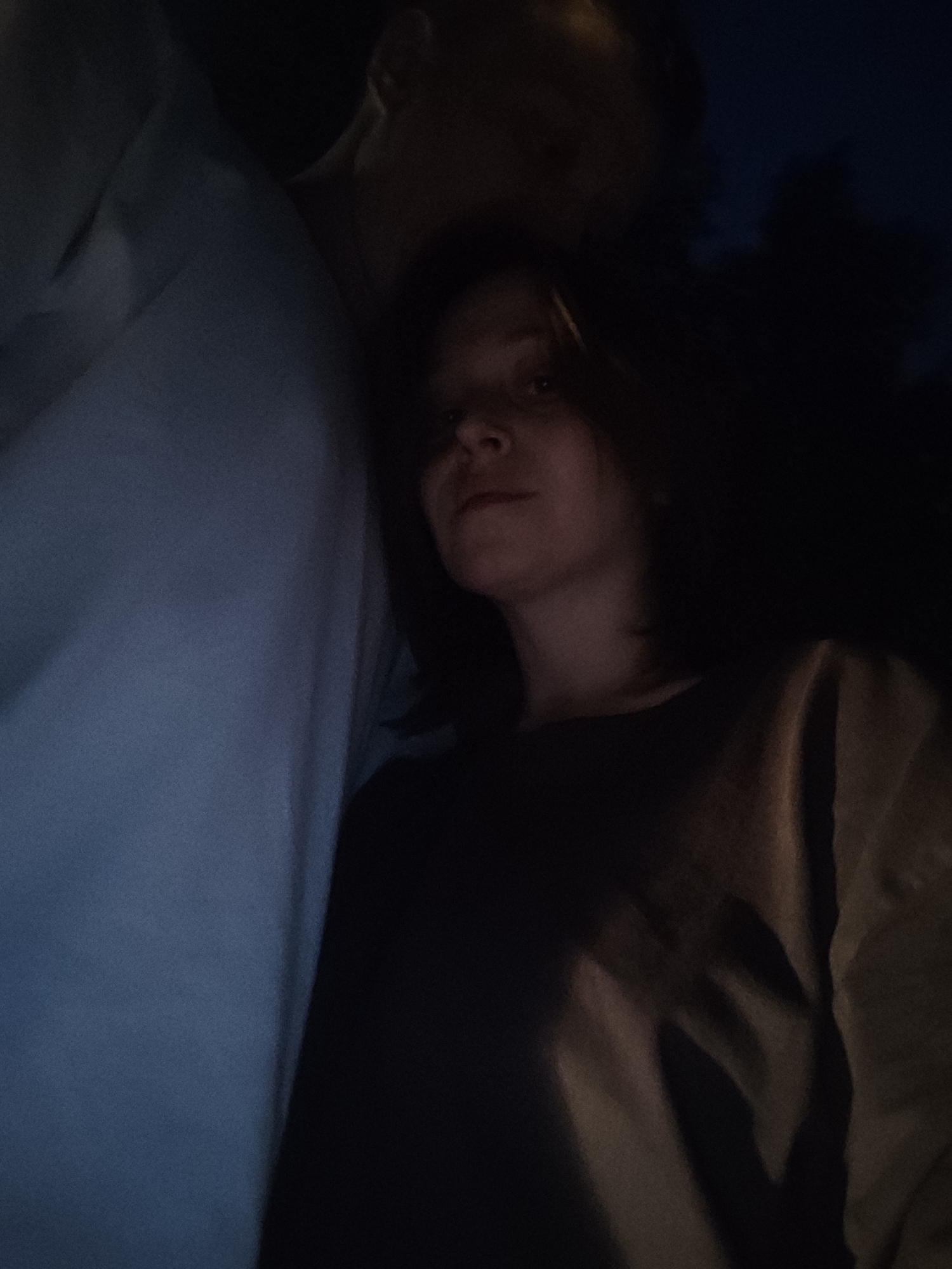 The image is a selfie taken at night, outdoors, in the city of Chaikovsky, Russia. Two people are partially visible. The person in the foreground is a young woman with dark hair, looking directly at the camera with a neutral expression. She is wearing a dark-colored top. Behind her, another person, whose face is obscured by shadow and being out of focus, appears to be standing close. The background is dark, suggesting it is nighttime with some natural light present. The overall lighting is dim and moody, with the faces of the subjects catching the available light. The location appears to be a public space given the context of a selfie.