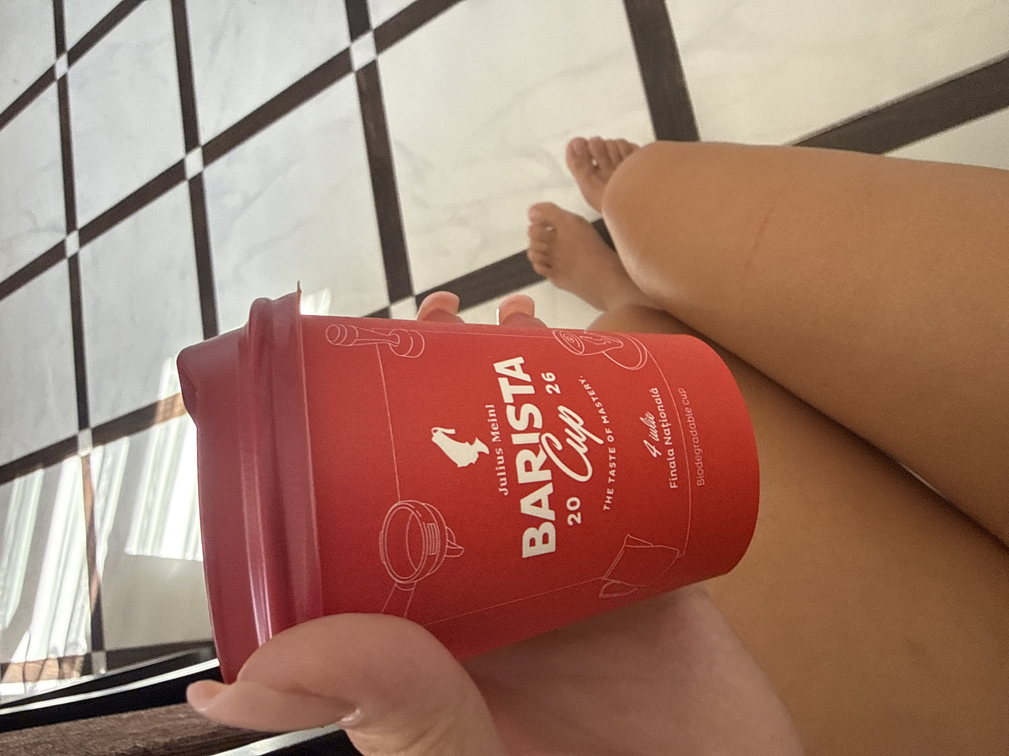 A close-up, overhead shot captures a person holding a bright red disposable coffee cup with a white lid. The cup features white text and illustrations related to coffee. Text on the cup reads "Julius Meinl," "BARISTA Cup," "20 26," and "THE TASTE OF MASTERY." Below this, it says "4 iulie Finala Națională" and "Biodegradable cup." The background is a tiled floor with a black and white checkered pattern. The lower half of the image shows the person's legs and feet, suggesting they are seated. The lighting indicates it is daytime. The image appears to have been taken indoors, potentially in a cafe or home setting. There is no specific indication of the city of Chișinău, Moldova in the image itself.