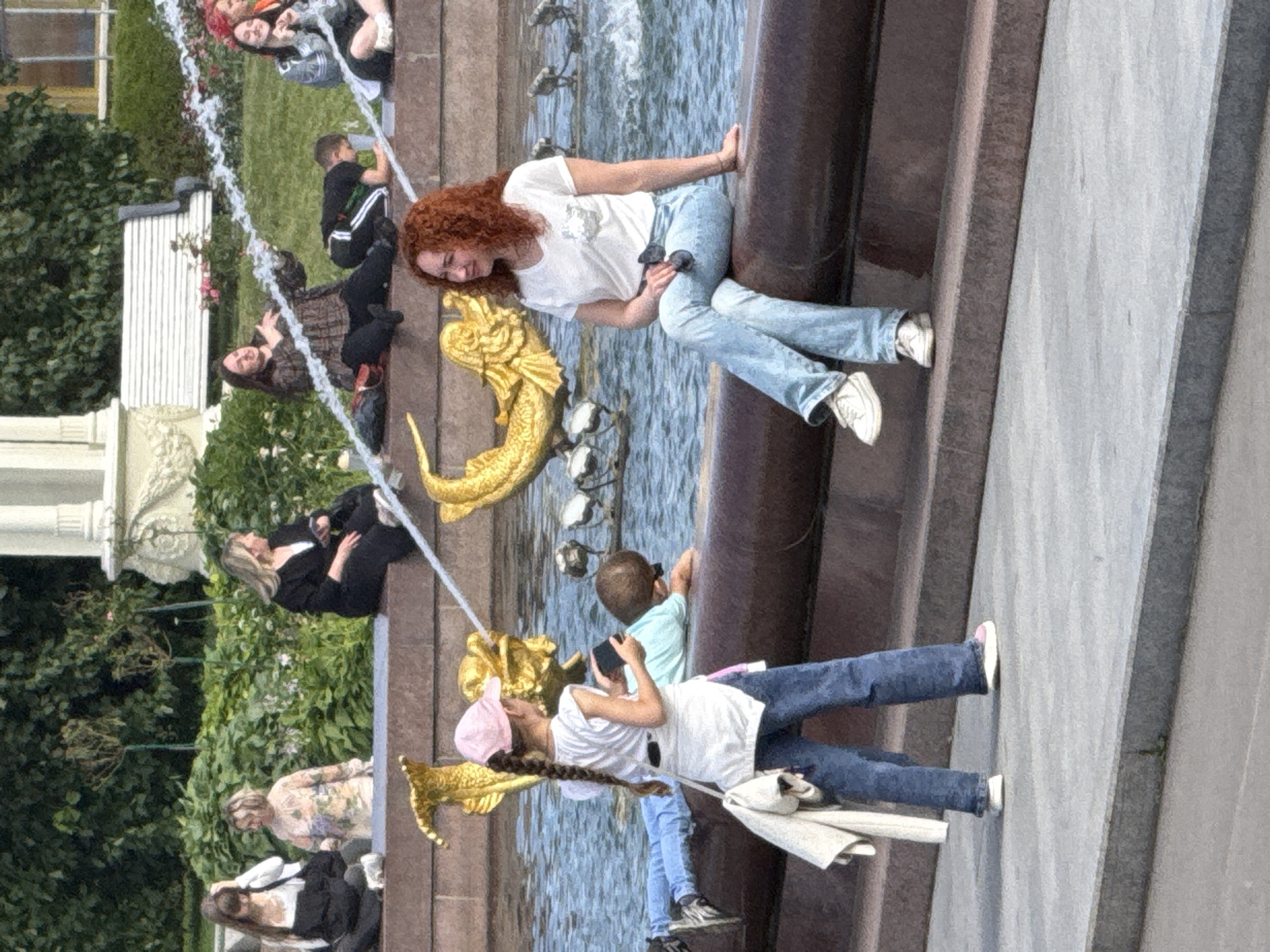 In this outdoor photograph, several people are gathered around a fountain adorned with gold sculptures. A young woman with curly red hair sits on the edge of the fountain, wearing a white t-shirt and light blue jeans. Next to her, another young woman with blonde hair is dressed in a black suit. A young boy in a light blue shirt is also present, seemingly holding a phone. Closer to the foreground, a child wearing a pink hat and a white shirt is holding a phone, while another child in a blue t-shirt is visible.
The scene appears to be a public space, likely a park or plaza, given the fountain and greenery. The water from the fountain is actively spraying. The time of day seems to be daytime, with natural light illuminating the scene. The overall atmosphere appears relaxed, with people enjoying the surroundings. There is no visible text in the image. The location is identified as Moscow, Russia.