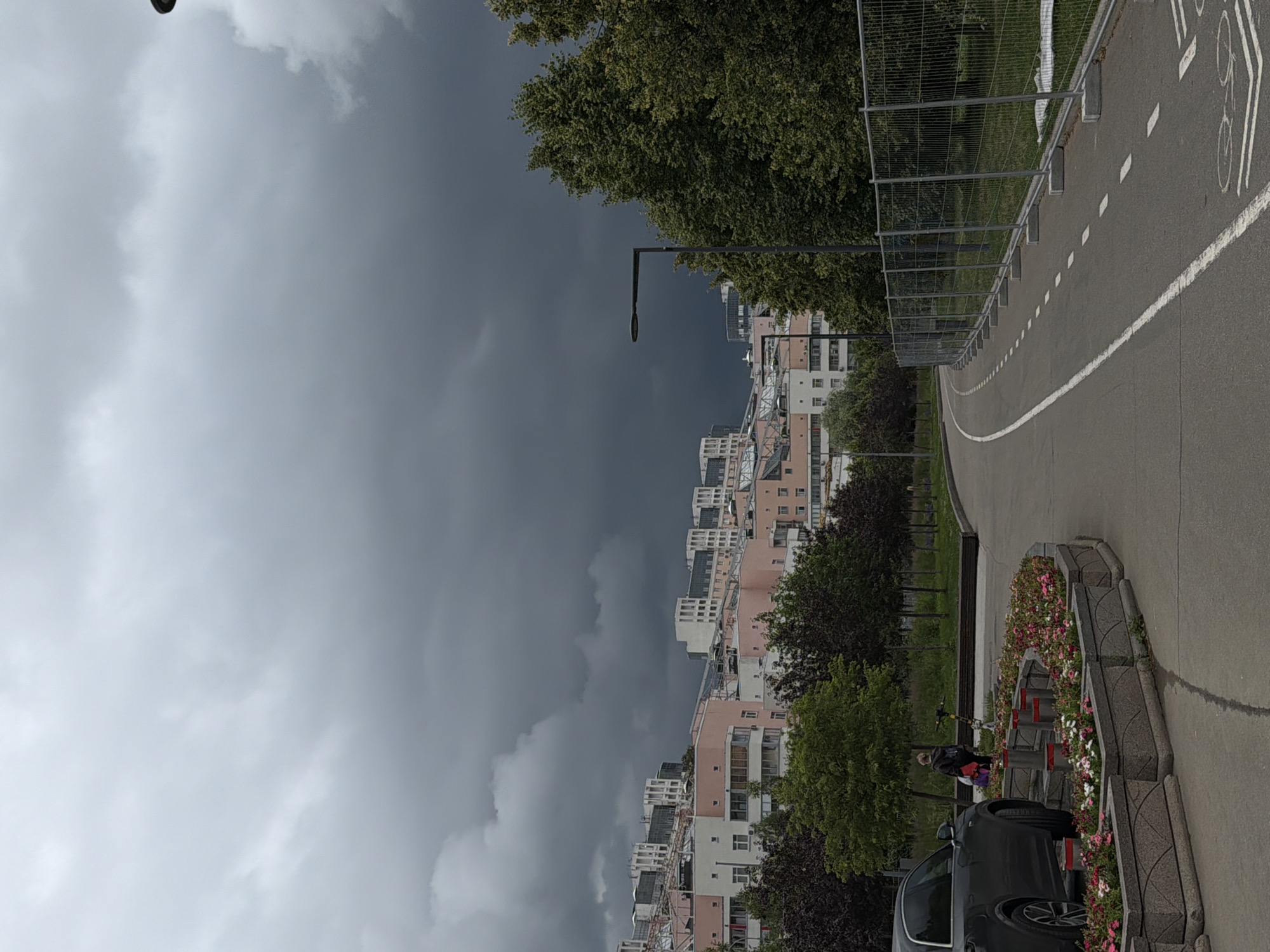 The image captures an outdoor scene with a dramatic sky and urban elements. A dense cluster of apartment buildings, characterized by their light pink and white facades, dominates the middle ground, interspersed with lush green trees. A dark, cloudy sky looms overhead, creating a stark contrast with the brighter, partly cloudy sky visible on the left.
In the foreground, a paved pathway, likely a bike lane indicated by a white line and bicycle symbol, curves to the right. Adjacent to the pathway is a decorative planter filled with colorful pink and white flowers, framed by grey stone borders. A dark grey car is parked near the planter, partially obscuring a person walking with a red bag.
The scene appears to be set in a city, possibly in the late afternoon or early evening, given the soft lighting and the ominous clouds that suggest an approaching storm. The mood is somewhat somber due to the weather, yet the presence of the residential buildings and recreational path hints at a normal day in a populated area. No visible text is present in the image.