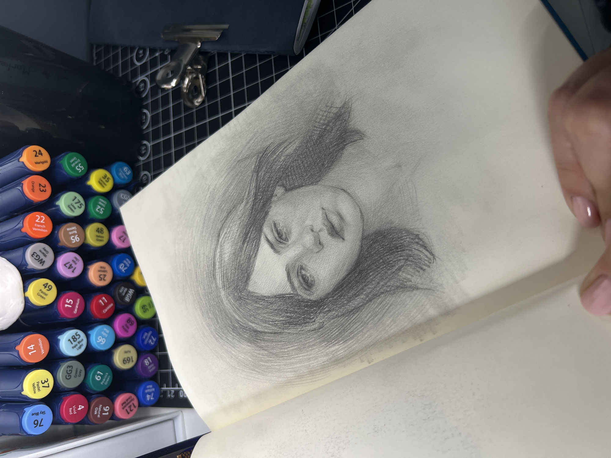 A pencil sketch of a woman's face is shown in an open sketchbook. The woman has dark, wavy hair and her head is tilted slightly to her right. She is looking directly at the viewer with a neutral expression. The sketch is rendered with a lot of detail, particularly in the eyes and lips. The background of the sketch is smudged, creating a soft halo effect around her head.
To the left of the sketchbook, a set of markers are organized in a white tray. The markers have colored caps, and some are labeled with numbers and color names. The markers appear to be for art use, such as Copic markers, given the branding visible on some caps.
In the background, a dark blue notebook or binder is partially visible, held in place by a silver binder clip. The entire scene is laid out on a black cutting mat with a grid pattern. The lighting is bright, casting subtle shadows. The image is taken from a high angle, looking down at the sketchbook and art supplies. There are no indications of a city or specific location.