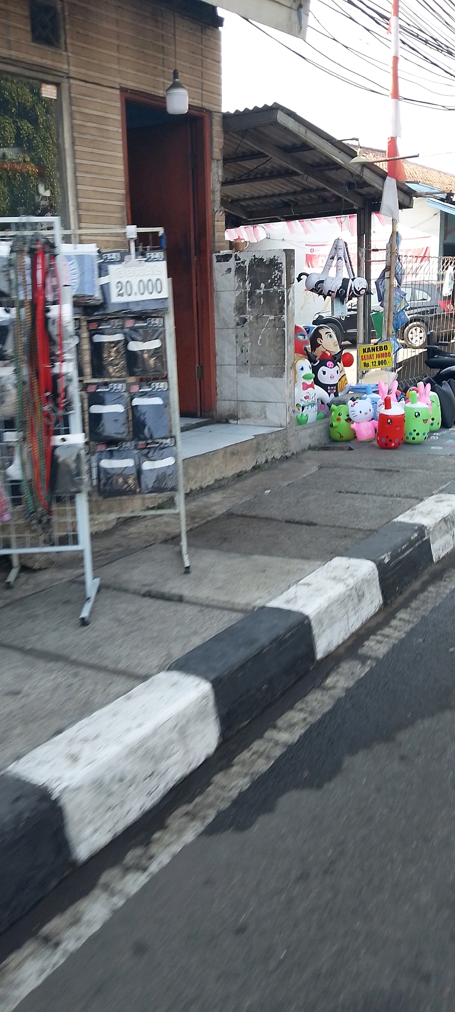 In Depok, Indonesia, a street scene unfolds with a sidewalk and a road demarcated by a concrete barrier painted with alternating black and white stripes. On the sidewalk, a metal rack displays various items, possibly accessories, with a sign indicating a price of "20.000". Next to the rack, several plush toys are arranged, including cartoon characters and animal figures, some resembling bubble tea. Further down the sidewalk, a red door leads into a building with tiled walls. Overhead, power lines crisscross the sky, and a flag with red and white stripes is visible. A parked vehicle and a portion of a building with a corrugated metal roof are also present. The overall impression is of a bustling commercial area with goods displayed for sale on the sidewalk.