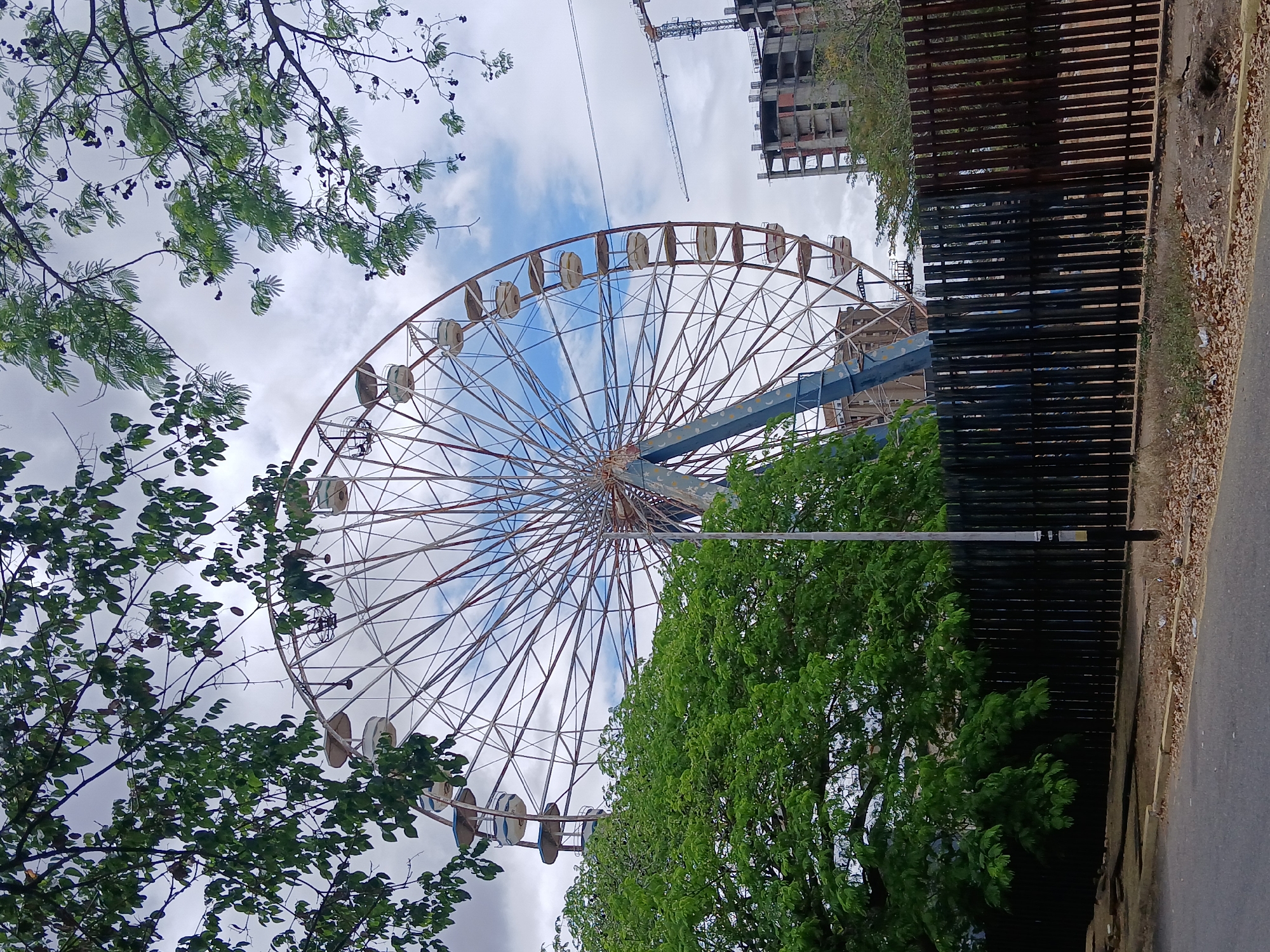 The image shows a large Ferris wheel in Ciudad Guayana, Venezuela. The wheel is partially obscured by trees in the foreground. A building under construction, along with a crane, is visible in the background. A black fence borders the right side of the image, alongside a paved road. The sky is cloudy with patches of blue.