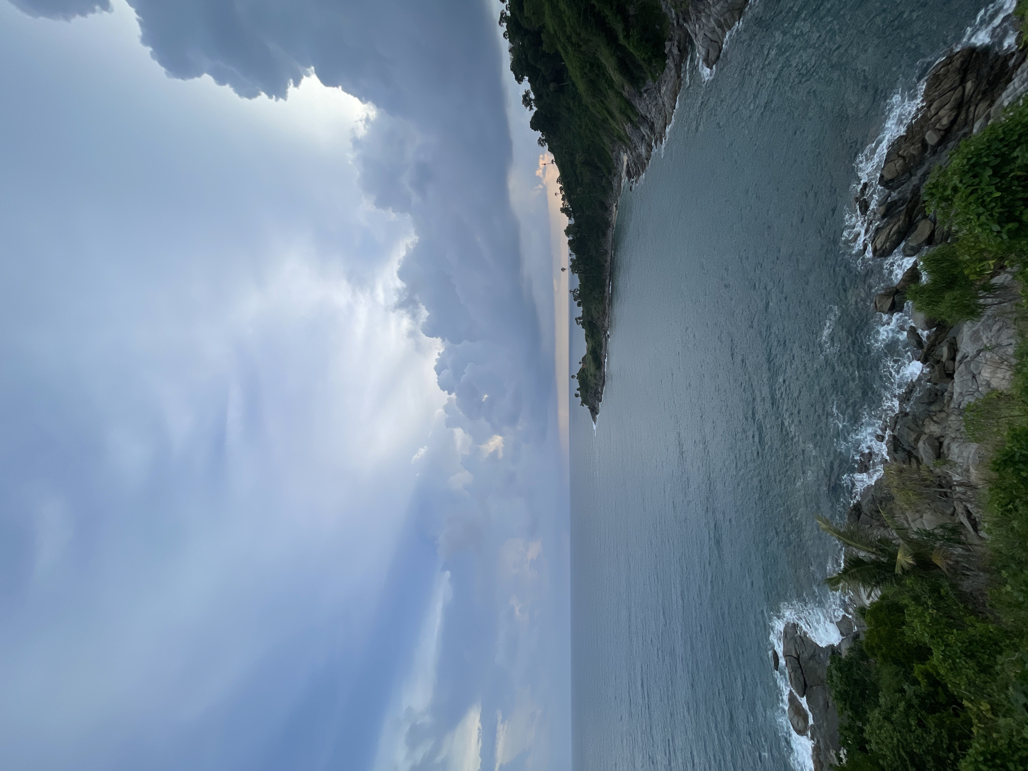 The image shows a scenic view of a coastline in Rawai, Thailand. The scene is dominated by a vast expanse of the ocean, with waves gently crashing against rocky shores. Lush green vegetation covers the hills that slope down to the water's edge. In the distance, a small peninsula juts out into the sea, dotted with trees. The sky above is a mix of blue and dramatic clouds, suggesting either the approach of a storm or the fading light of dusk. The overall impression is one of natural beauty and tranquility.