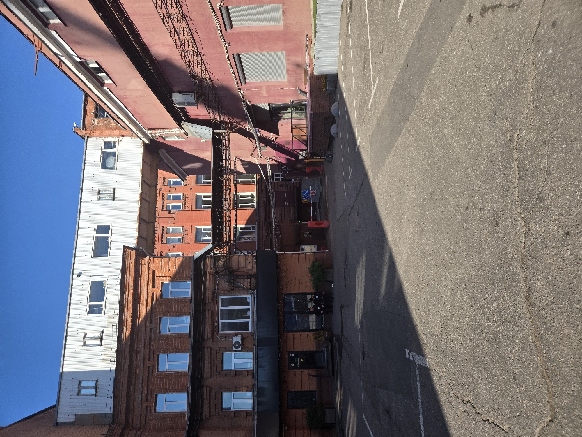 The image shows an outdoor courtyard area in Moscow, Russia, likely a commercial or industrial space, with several brick and metal buildings. In the foreground, cracked asphalt covers the ground, marked with white lines indicating parking spaces. A large shadow from a building falls across the courtyard.
In the mid-ground, a two-story brick building with multiple windows is visible. The ground floor of this building appears to be a commercial establishment with a dark entrance and signage. Two individuals wearing dark uniforms, possibly security guards, are seated on a ledge in front of this building. To the right of the brick building is a larger, red-colored industrial-style building with roller-shuttered windows and visible external wiring and conduits. This building casts a long shadow.
Further back, a modern-looking white metal structure is situated on top of another section of the brick building, creating a layered architectural effect. The background is dominated by a clear blue sky. The scene appears to be captured during daylight hours, with strong sunlight creating distinct shadows. An automated barrier gate is visible in the distance, suggesting access control to the area.