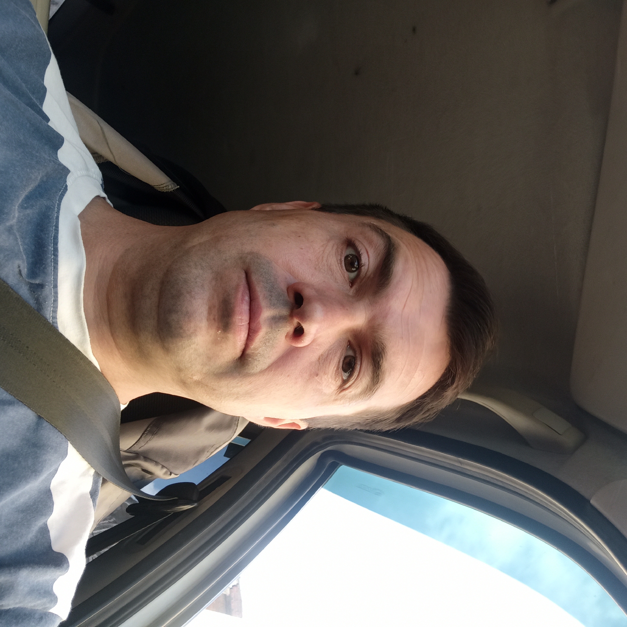 In a frontal, eye-level selfie taken from inside a vehicle, a man with fair skin, short brown hair, and a light stubble is the primary subject. He is looking directly at the camera with a neutral expression. His features include brown eyes, a straight nose, and closed lips. He is wearing a blue and white striped t-shirt and a dark seatbelt is visible across his chest. The background of the image shows the interior of a car, including the headliner and a portion of the open car door with a view of a bright sky. The lighting suggests it is daytime. This scene is not specific to Nizhny Novgorod, Russia, as there are no identifiable landmarks or features to indicate a particular location within the city.