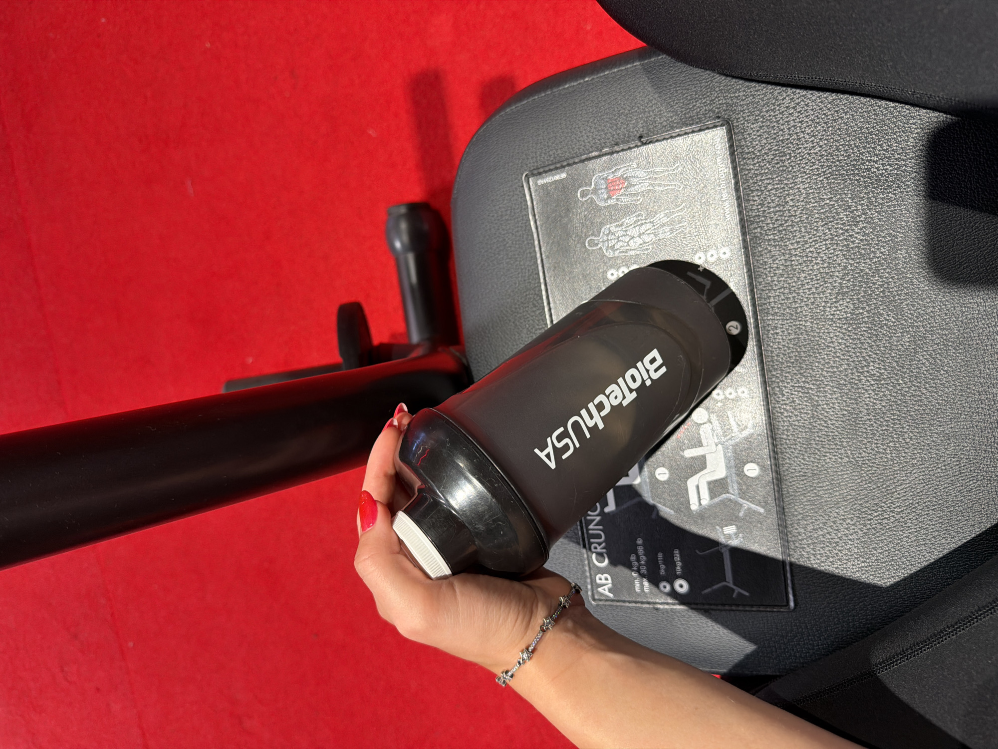 A person's hand with red nail polish is holding a black and white shaker bottle with the logo "BiotechUSA" on it. The shaker is positioned over a black leather seat with instructions for an "AB CRUNCH" exercise machine visible. The seat is part of gym equipment. The background is a bright red surface, likely a gym floor. The image appears to be taken in a fitness center. There is no indication this image is related to the city of Strasbourg, France.