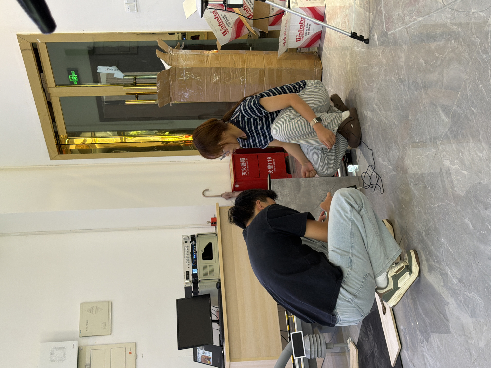 In a well-lit indoor space, possibly a storage area or workshop, two individuals are actively engaged in what appears to be a construction or assembly task. The room features a smooth, light-colored, marble-patterned floor. To the left, a light-colored desk holds a computer monitor, a laptop, and some electronic equipment, suggesting a workspace.
A man, dressed in a dark t-shirt and light blue jeans, is crouched down on the floor, his back to the viewer. He is wearing a pair of green and white sneakers. He seems to be working on something on the floor, possibly positioning or securing a large, flat, light-colored object, perhaps a tile or a panel.
A woman, wearing a striped t-shirt and striped trousers, is also crouched down, facing the man. She has her hair tied back and is wearing glasses. She appears to be assisting the man, reaching towards the same object on the floor. She is wearing brown clogs.
In the background, several cardboard boxes are stacked, some of them open, containing items that might be related to the task. One box prominently displays the "Wahaha" brand, suggesting it contains beverages. A red fire extinguisher cabinet is also visible, with Chinese text indicating "FIRE EXTINGUISHER." A double door with gold trim and glass panels is to the right, with a green "EXIT" sign illuminated above it. The overall environment suggests a functional space where unpacking, assembly, or installation is taking place. The presence of the "Wahaha" brand might suggest this location is in or around the city of Jiangjin, China, as Wahaha is a prominent Chinese company.