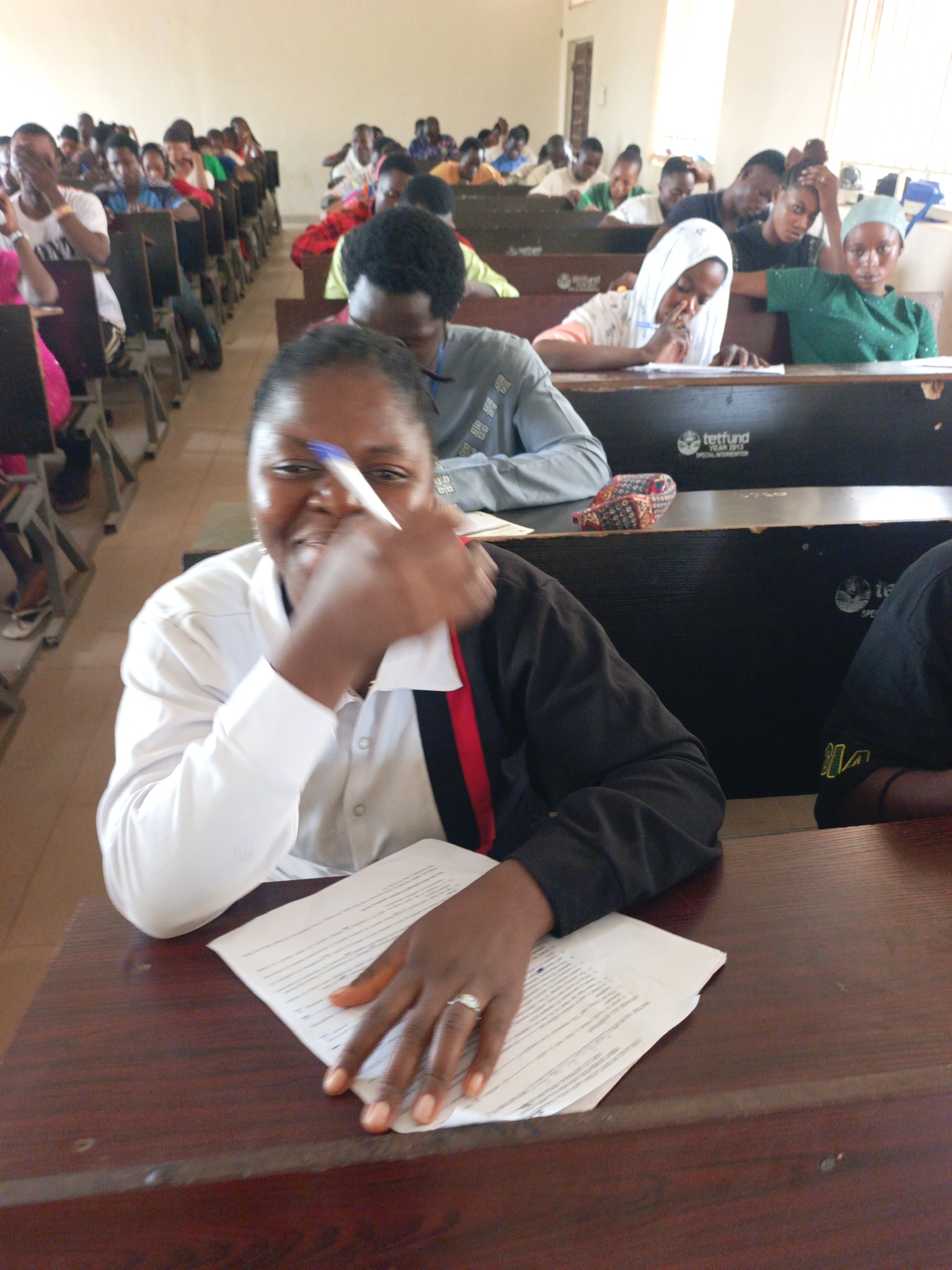The photo, likely taken in Akwanga, Nigeria, features a university classroom filled with students taking an exam. In the foreground, a woman in a white and black top smiles, holding a pen. A sheet of paper with text lies on the wooden desk in front of her. Other students are seated at rows of desks behind her, some writing, some looking down at their papers, and others gazing forward. The desks have the inscription "tetfund YEAR 2013 SPECIAL INTERVENTION." The walls are painted in a light, neutral color.