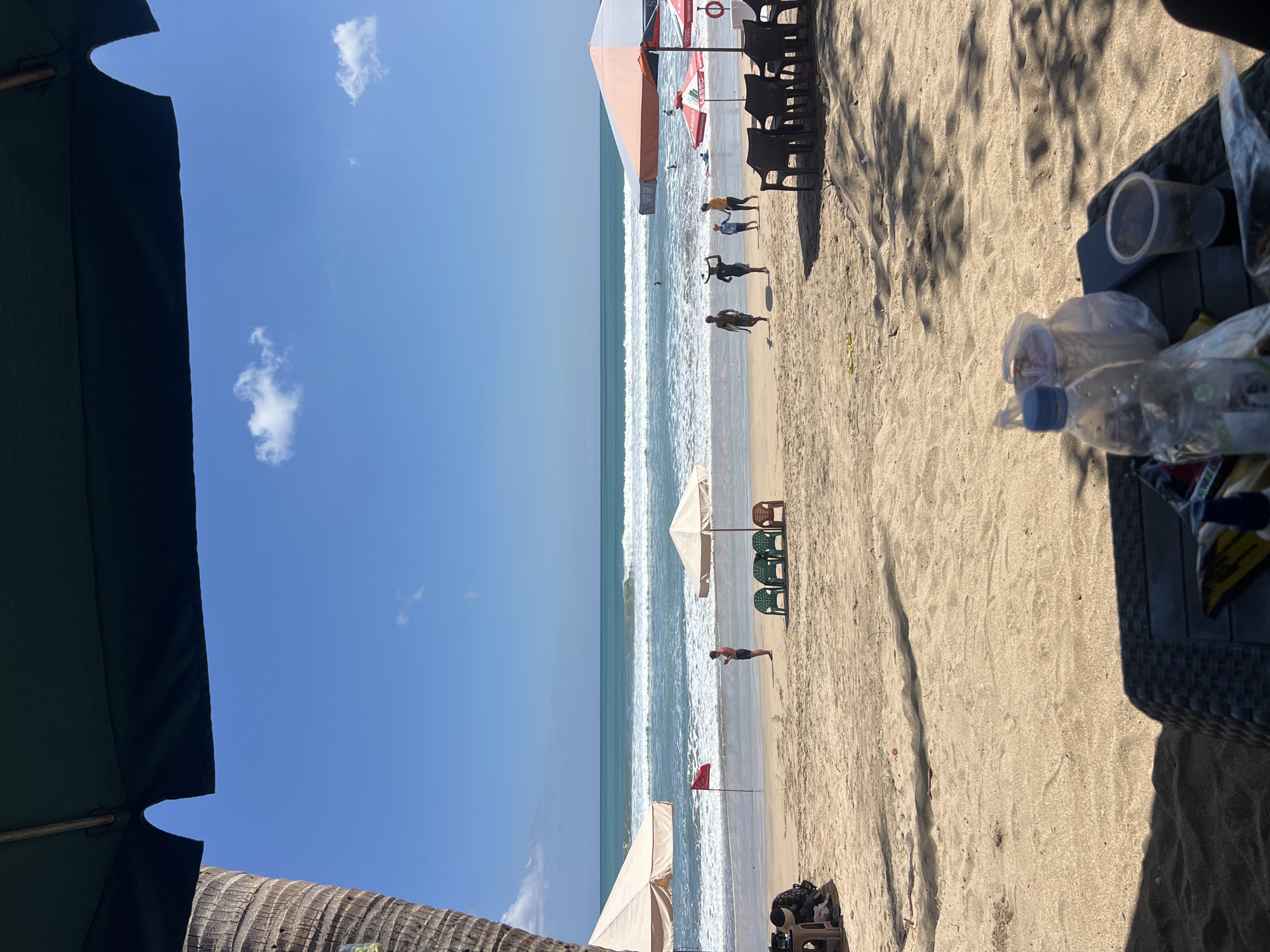 In Kuta, Indonesia, a sunny beach scene unfolds under a clear blue sky with scattered white clouds. The foreground shows a dark woven table with a plastic bottle of water, two plastic cups, a smartphone, and other items, casting shadows on the sandy ground. Behind the table, the beach stretches towards the ocean. Several beach chairs, some green and some brown, are arranged near an open umbrella.
Further down the beach, a red flag is planted in the sand. Several people are visible in the distance. Some are walking along the shoreline, while others are in the ocean, with waves breaking. Two large umbrellas, one cream-colored and another with orange and white stripes, provide shade. A palm tree trunk is visible on the left side of the image, partially framing the scene. The ocean is a vibrant blue-green, with white foam from the waves. The overall atmosphere is one of leisure and activity on a tropical beach.