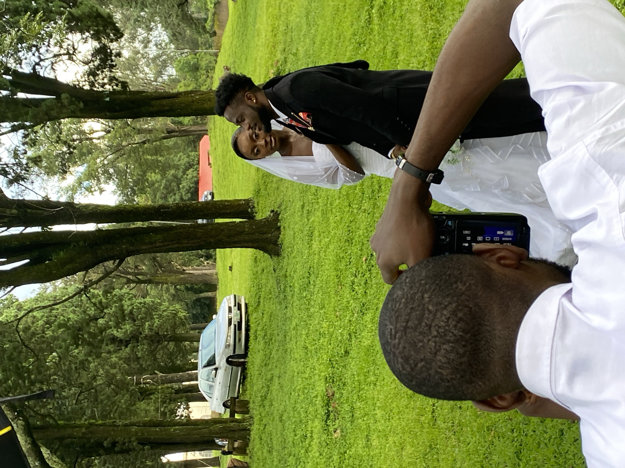 A wedding photoshoot is taking place on a lush green lawn in the city of Jos, Nigeria. A male photographer, wearing a white shirt, is in the foreground, holding a camera and focusing on the wedding couple. The bride, adorned in a white wedding gown and veil, stands next to the groom, who is dressed in a black suit with a patterned tie. Behind them, several tall trees line the background, with a silver sedan parked on a paved area to the left. The overall atmosphere appears to be a serene outdoor event.