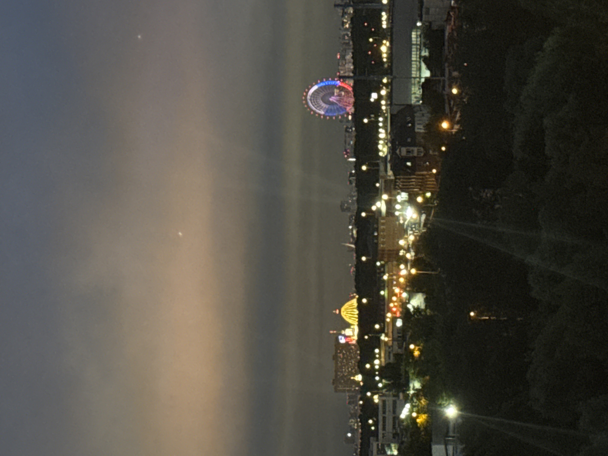 The image captures a nighttime view of Moscow, Russia, with a prominent Ferris wheel illuminated in red, white, and blue. In the background, a large, brightly lit domed structure, likely a landmark, is visible. The cityscape is dotted with numerous lights from buildings and street lamps. The sky above is a gradient of dark blue and a lighter, hazy orange, possibly due to light pollution or atmospheric conditions. The foreground is dominated by dark silhouettes of trees and some buildings, with streaks of light suggesting a long exposure or movement within the frame. The scene conveys a sense of urban evening ambiance.