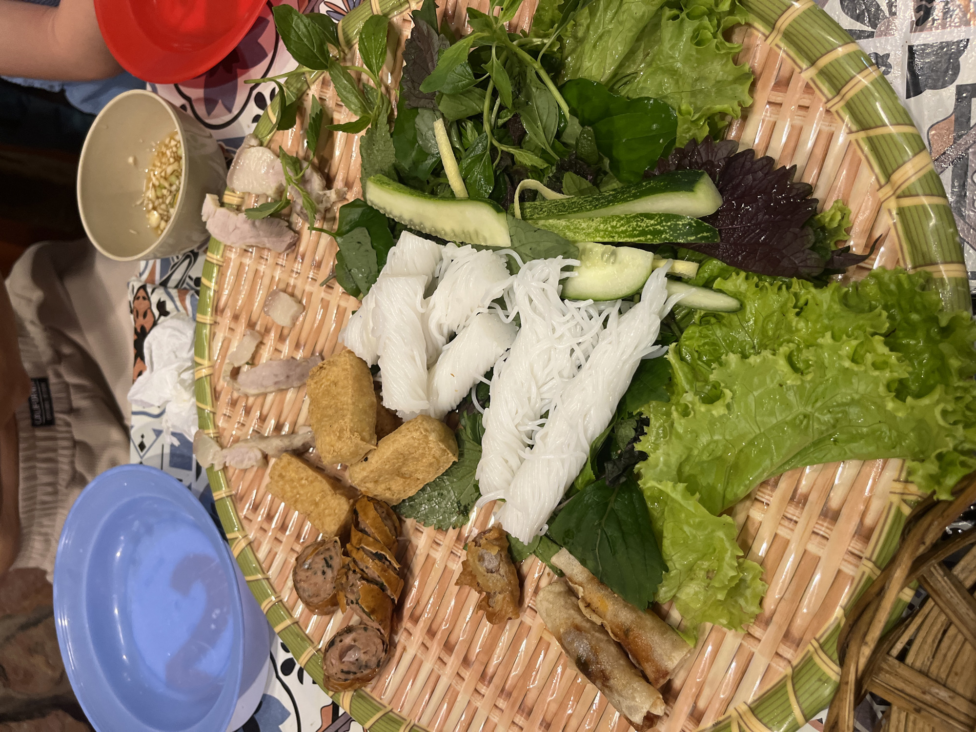 A platter of Vietnamese food is arranged on a woven bamboo tray. The platter includes thinly sliced cooked pork, fried tofu cubes, fried spring rolls, sliced sausage, rice vermicelli noodles, and a variety of fresh herbs and lettuce. A small bowl containing a dipping sauce with chopped scallions and what appears to be garlic is visible in the upper right portion of the image. There is also a blue bowl of water to the left and a red bowl on the far right. The food is laid out on a table with a patterned tablecloth. The scene suggests a meal is being served or prepared.